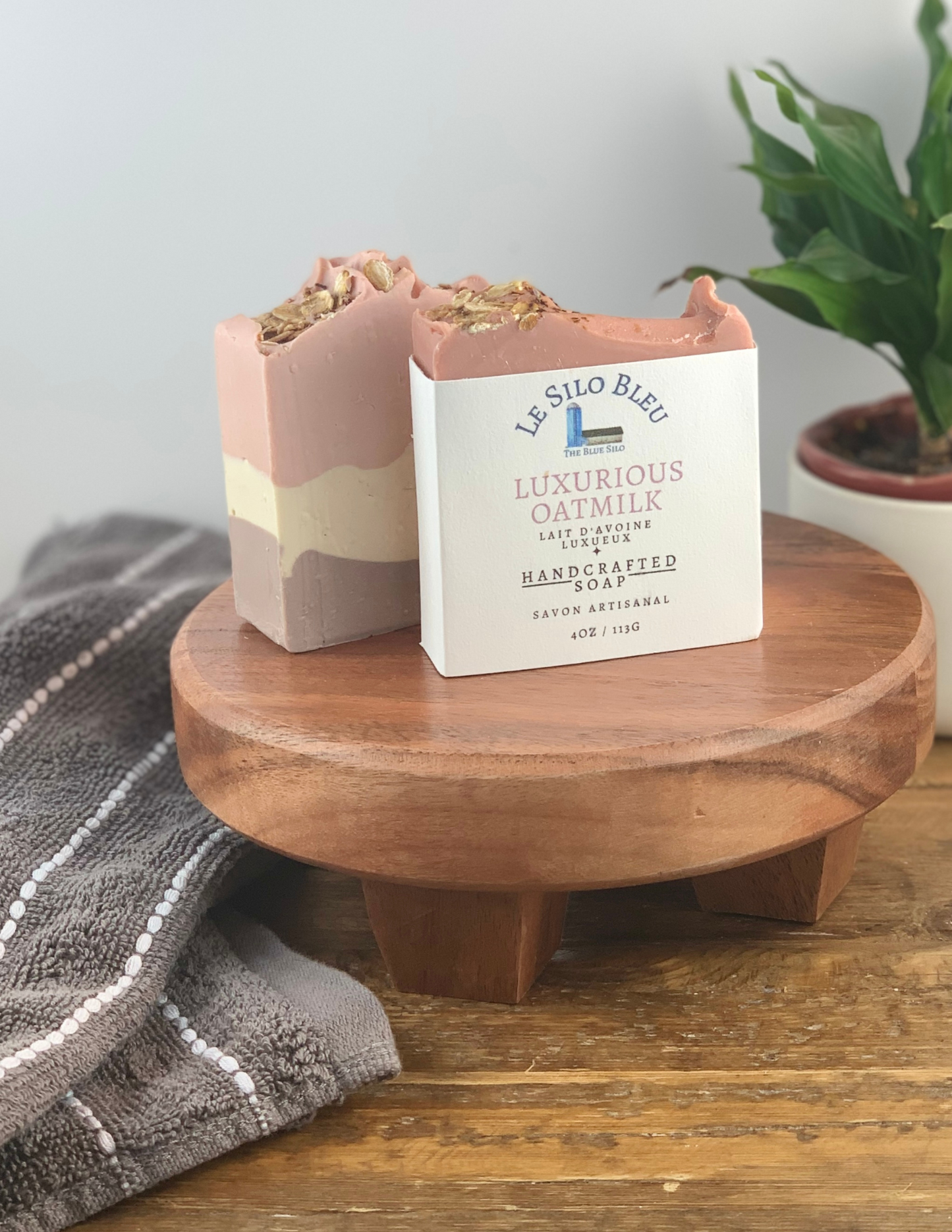 Two bars of Luxurious Oatmilk Soap sitting on a wooden platform with one soap wrapped in white labeling and one without. There is a green plant in a white pot in the background and a grey and white striped towel in the foreground. There is a wooden table and white background. 