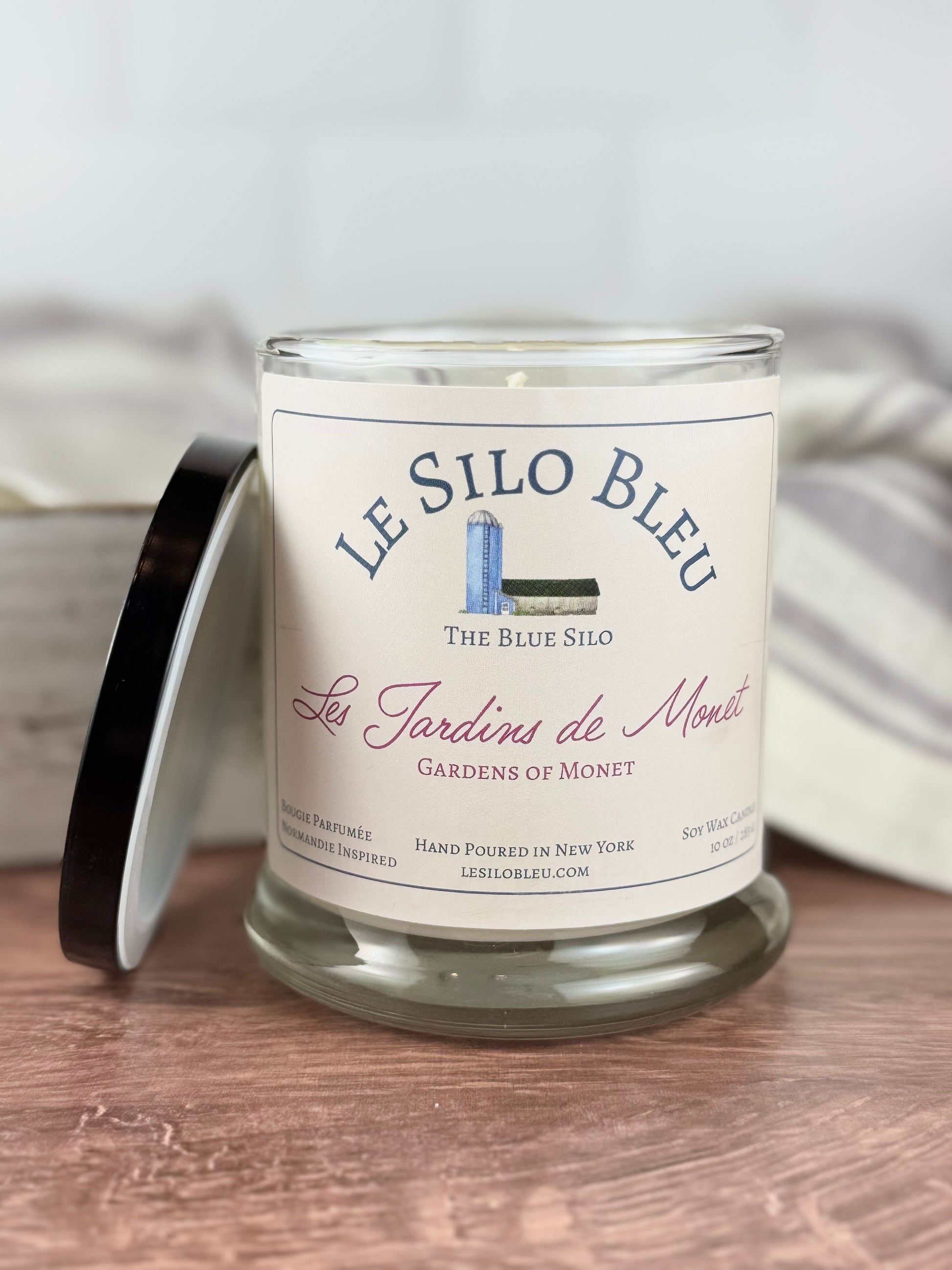 A large hand poured unlit soy wax candle in a glass jar labeled "Les Jardins de Monet - Gardens of Monet" sitting on a wooden tabletop with a black lid leaning against the jar. In the background you can see a white linen towel with lavender stripes in an antique box.