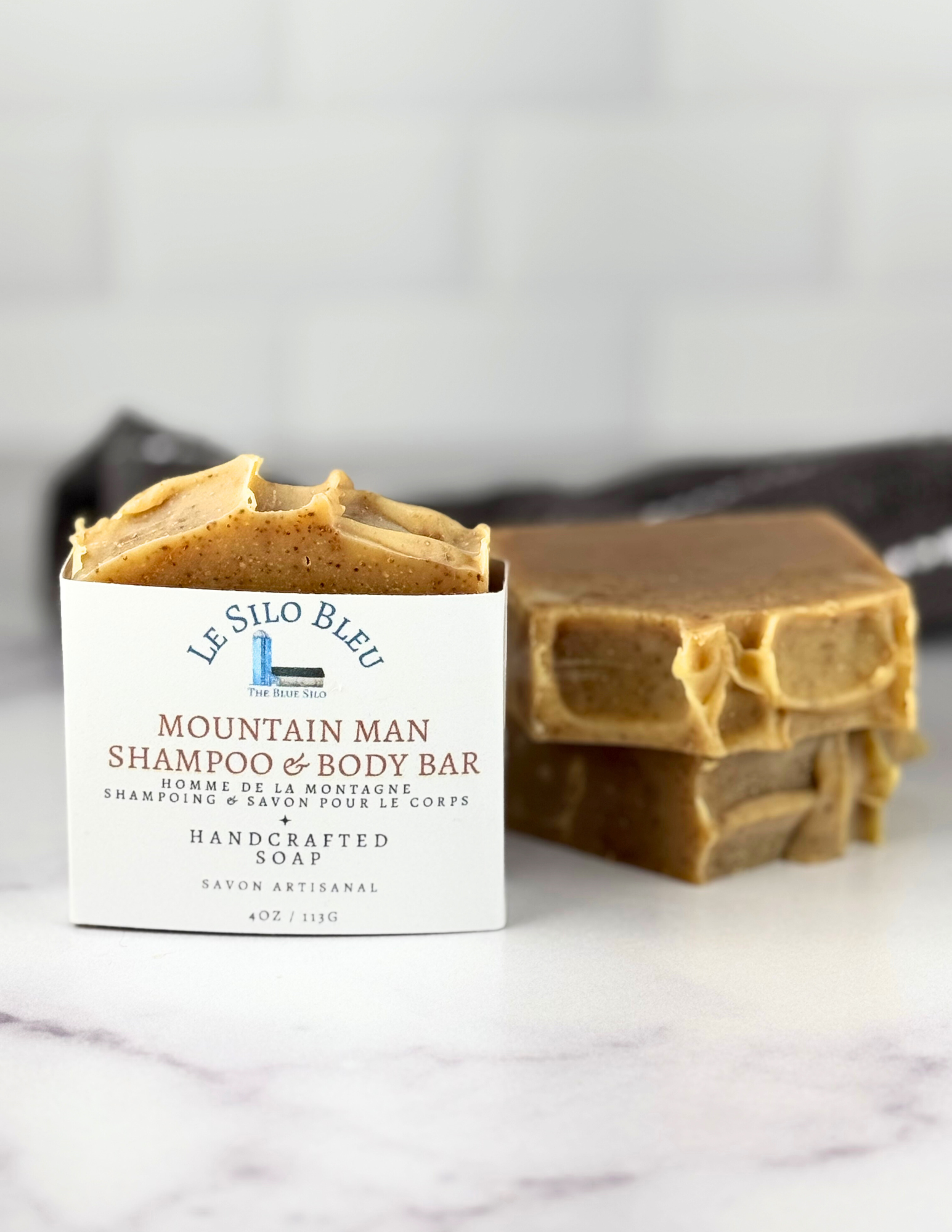 Stack of Mountain Man exfoliating soap on a white marble table with a grey striped towel in the back ground