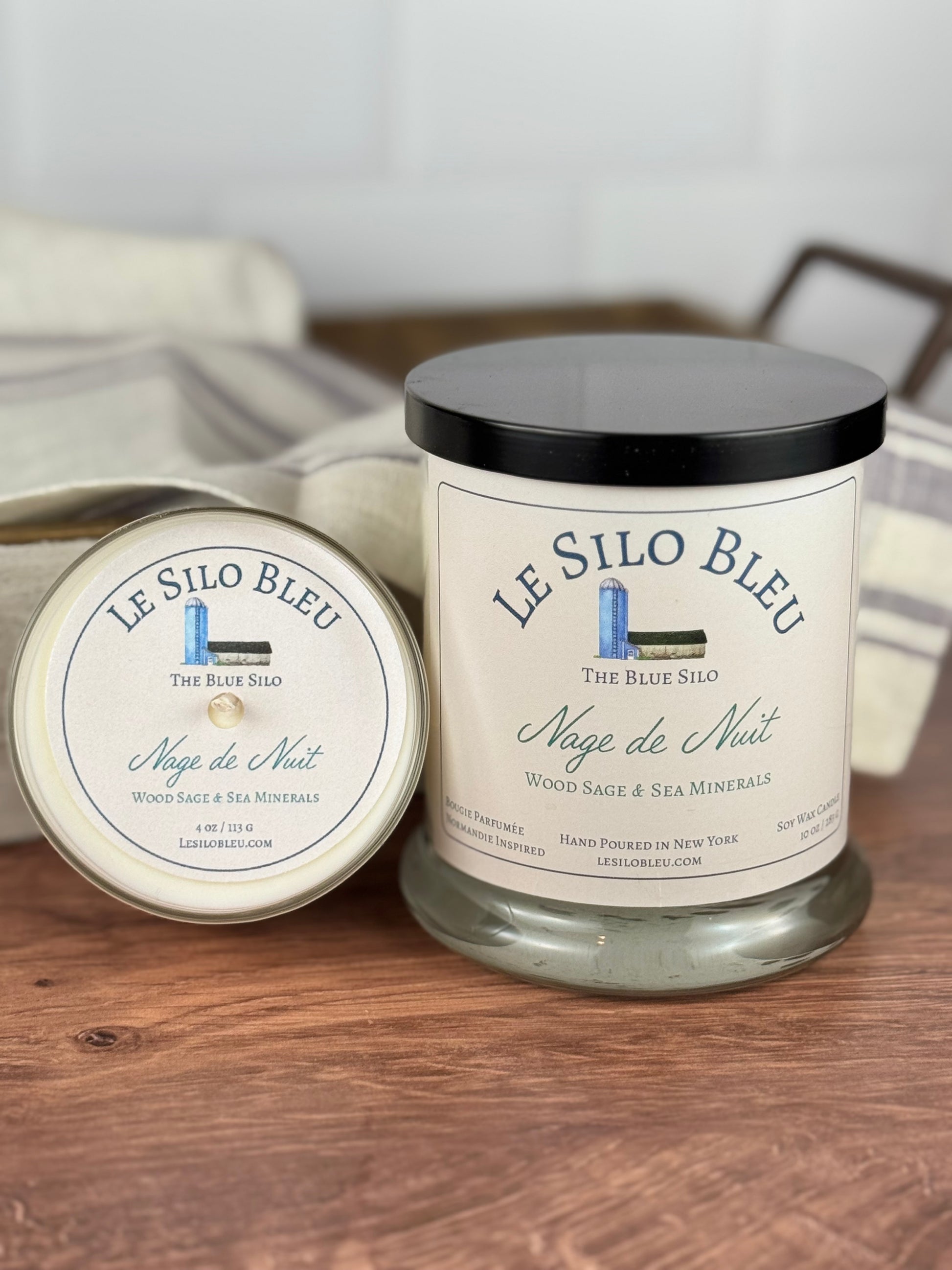 A large and a small hand poured unlit soy wax candle in glass jars labeled "Nage de Nuit-Wood Sage and Sea Minerals" sitting on a wooden tabletop with a black lid on the large jar. In the background there is a white linen towel with lavender stripes in an antique box.