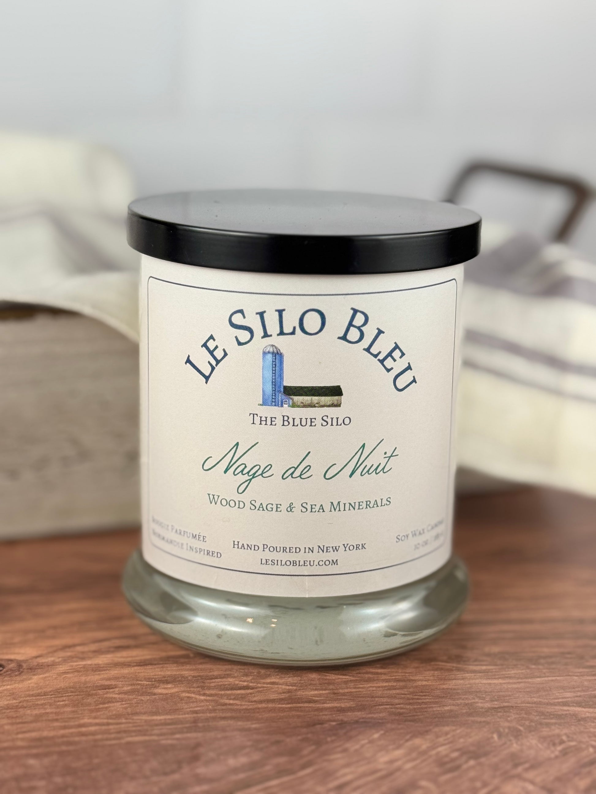 A large hand poured unlit soy wax candle in a glass jar labeled "Nage de Nuit-Wood Sage and Sea Minerals" sitting on a wooden tabletop with a black lid on the jar. In the background there is a white linen towel with lavender stripes in an antique box.