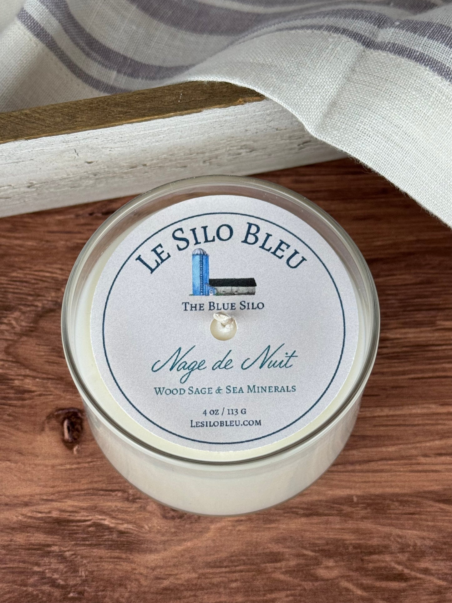 A small hand poured unlit soy wax candle in a glass jar labeled "Nage de Nuit-Wood Sage and Sea Minerals" sitting on a wooden tabletop. In the background there is a white linen towel with lavender stripes in an antique box.