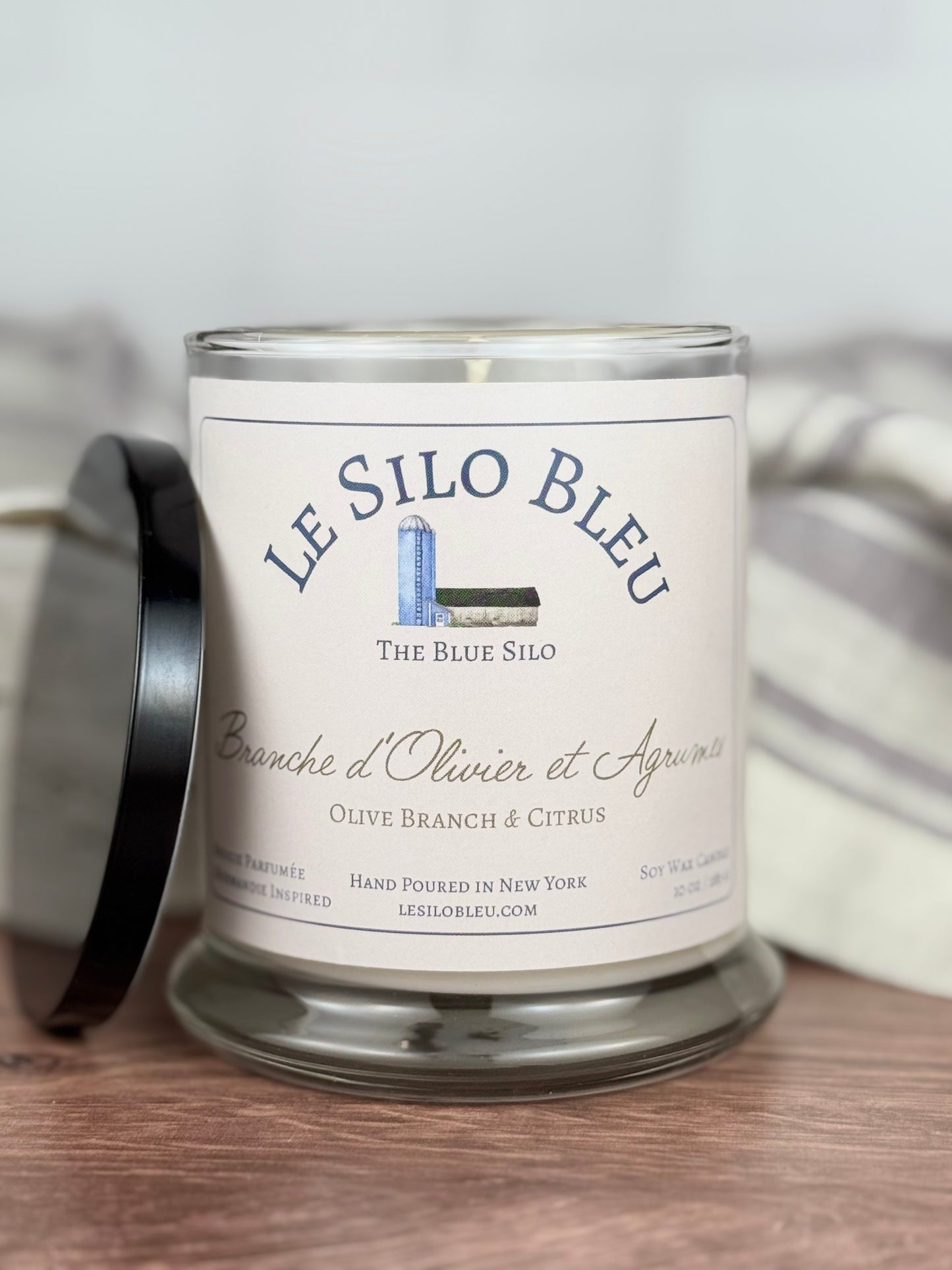 A large handpoured unlit soy wax candle in a glass jar labeled "Branche d'Olivier et Agrumes- Olive Branch and Citrus" sitting on a wooden table top with a black lid next to the jar. In the background there is a white linen towel with lavender stripes in an antique box.