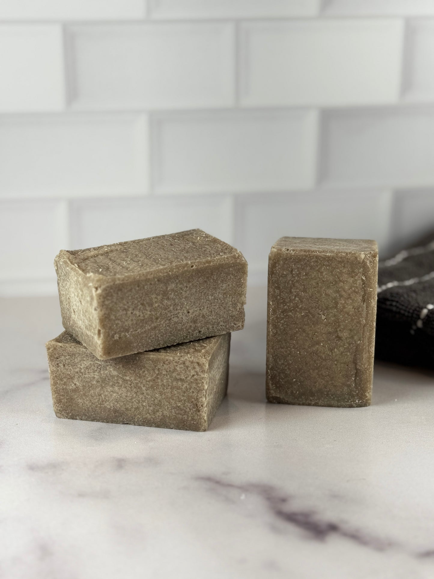 Three chunky bars of handmade sea salt soap in a muted taupe-brown color, arranged on a white marble surface with a white subway tile backsplash in the background.