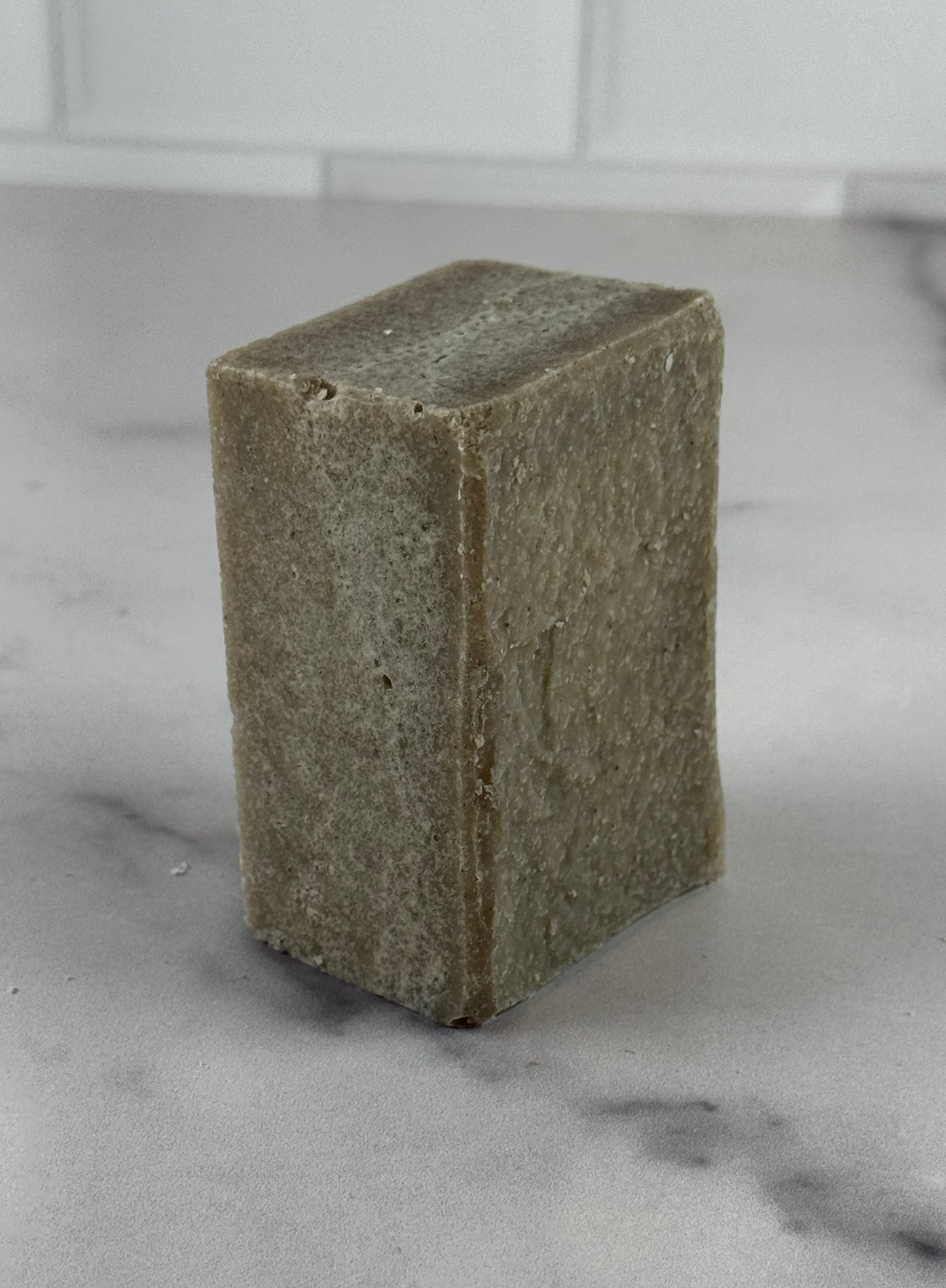 A chunky bar of handmade sea salt soap in a muted taupe-brown color, arranged on a white marble surface with a white subway tile backsplash in the background.