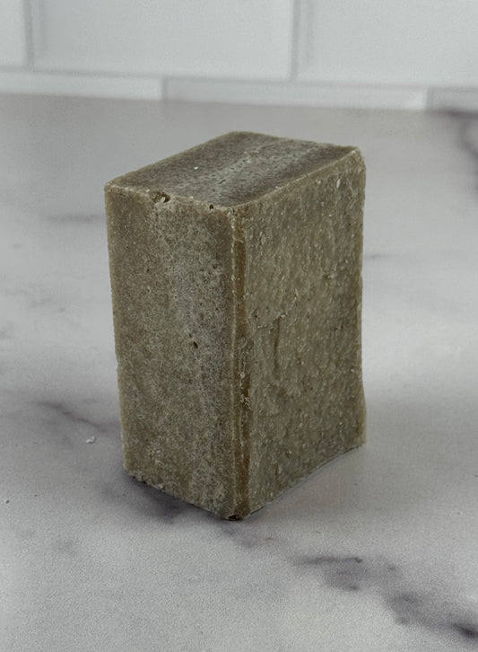 A chunky bar of handmade sea salt soap in a muted taupe-brown color, arranged on a white marble surface with a white subway tile backsplash in the background.