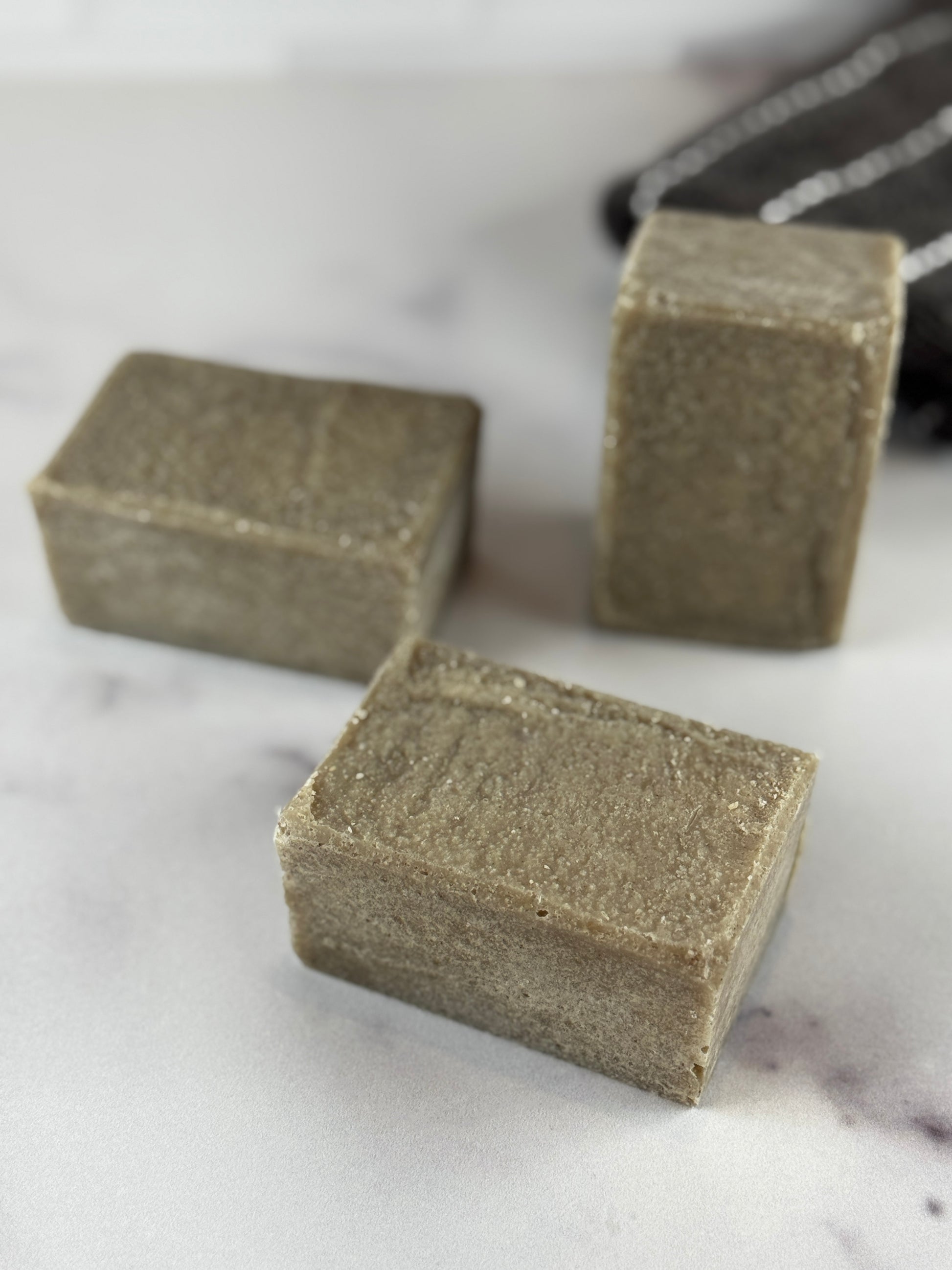 Three chunky bars of handmade sea salt soap in a muted taupe-brown color, arranged on a white marble surface with a white subway tile backsplash in the background.