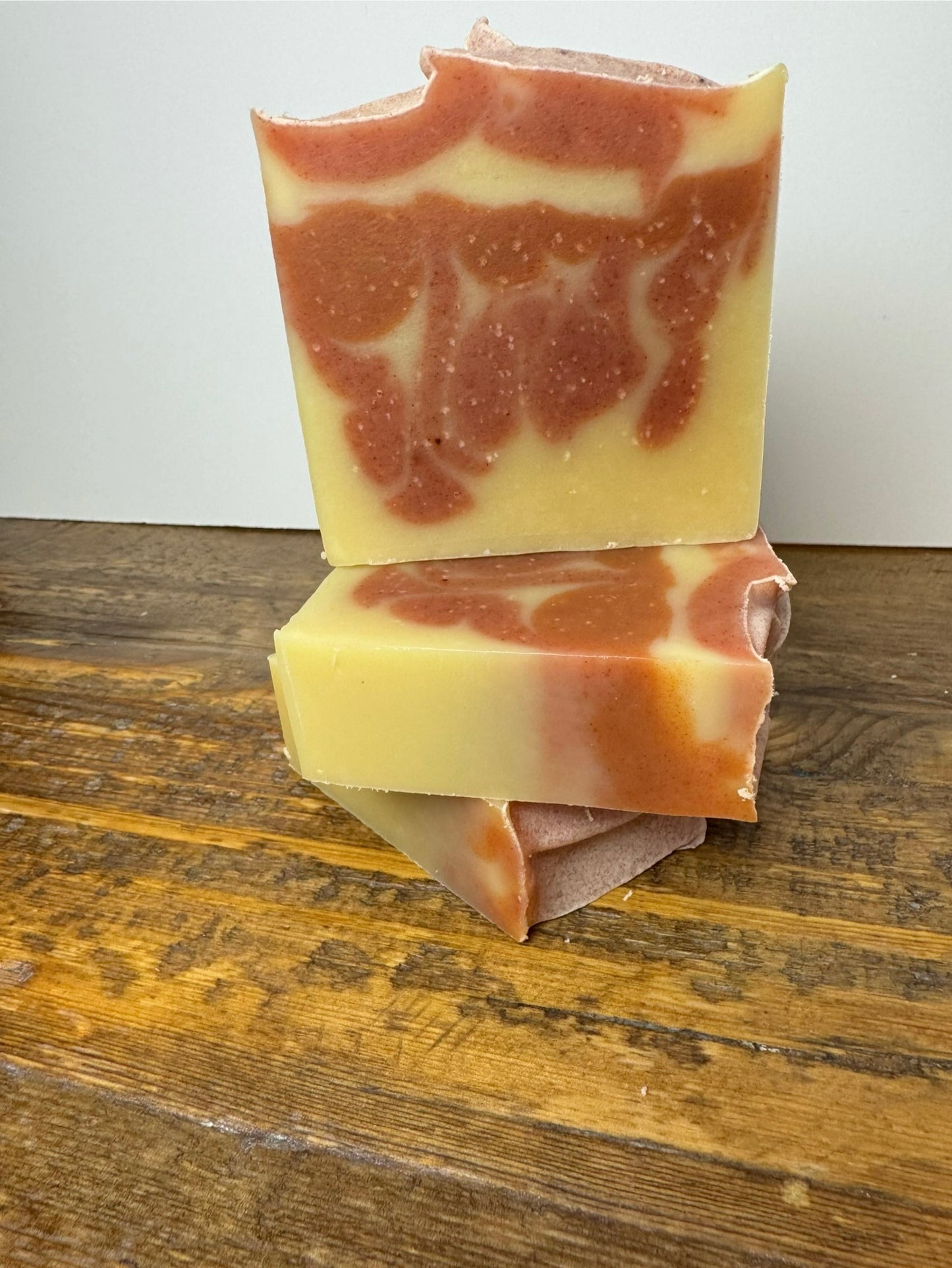 An all natural bar scented with Orange essential oil sitting on a wooden tabletop with a white background. This bar has orange swirls in a light yellow orange bar. 