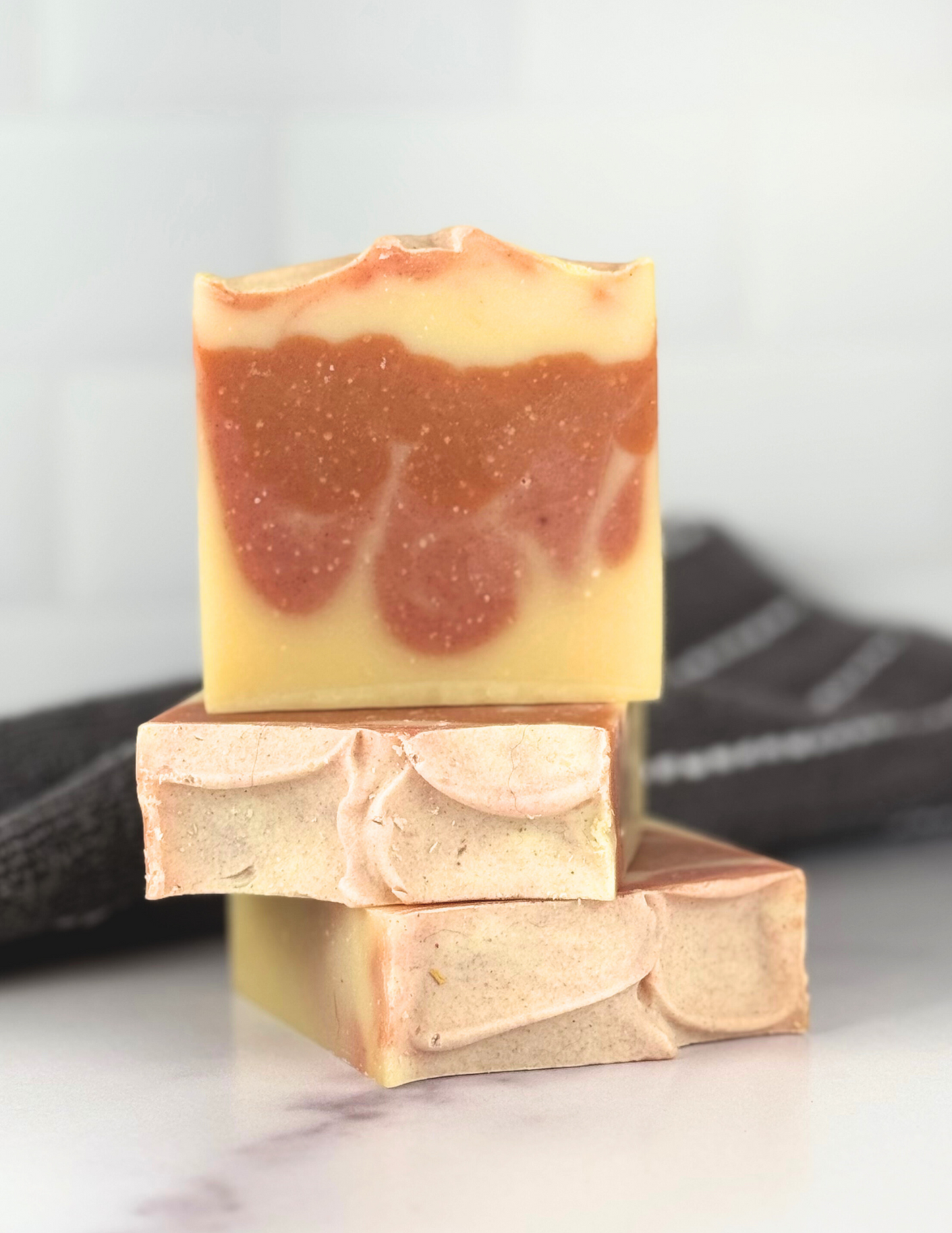 Three bars of Orange Blossom Soap sitting on a marble tabletop with a grey and white stripped towel in the background. The soap is two shades of orange and yellow swirled together. 