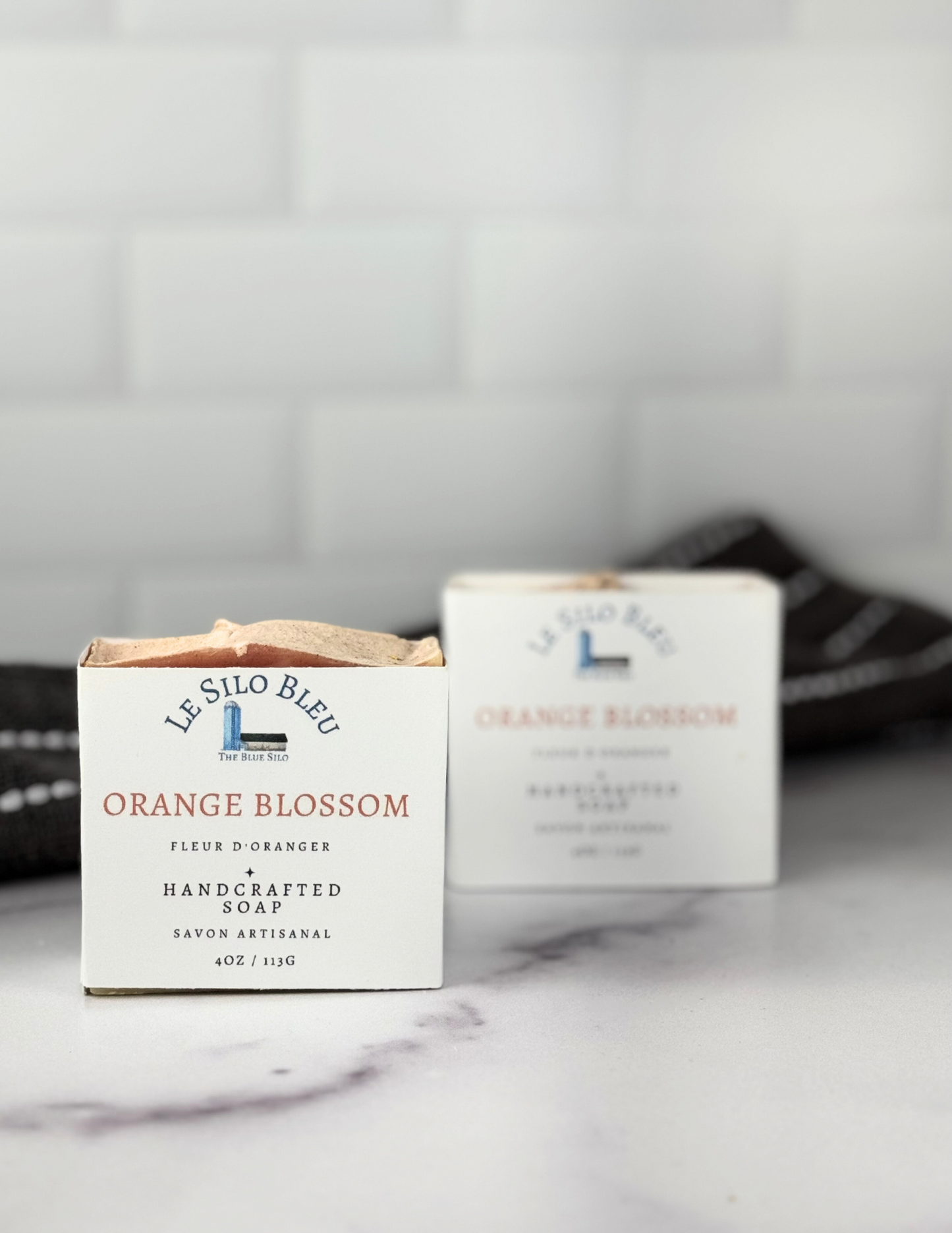 Two labeled bars of Orange Blossom Soap sitting on a marble tabletop with a grey and white stripped towel in the background. The soap label is white with our logo and soap name on it.