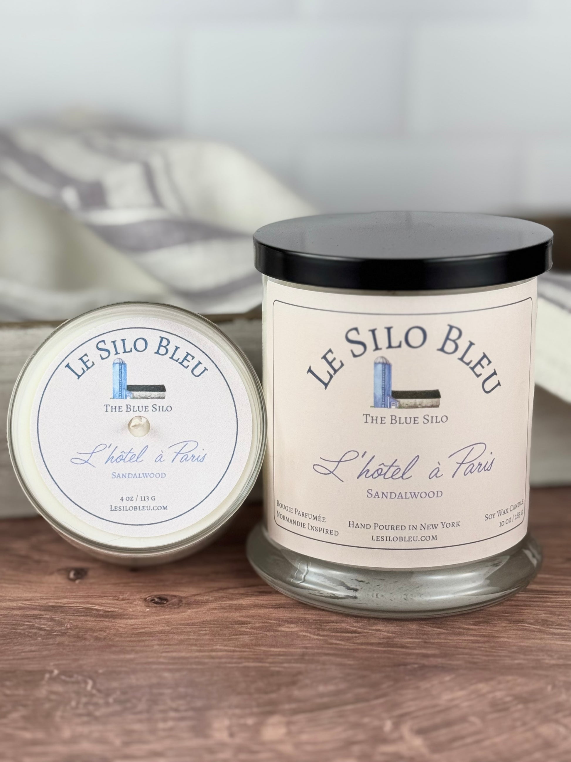 A large handpoured unlit soy wax candle in a glass jar labeled "L'hotel a Paris - Sandalwood" sitting on a wooden table top with a black lid on the large jar. In the background you can see a white linen towel with lavender stripes in an antique box with a metal handle. 