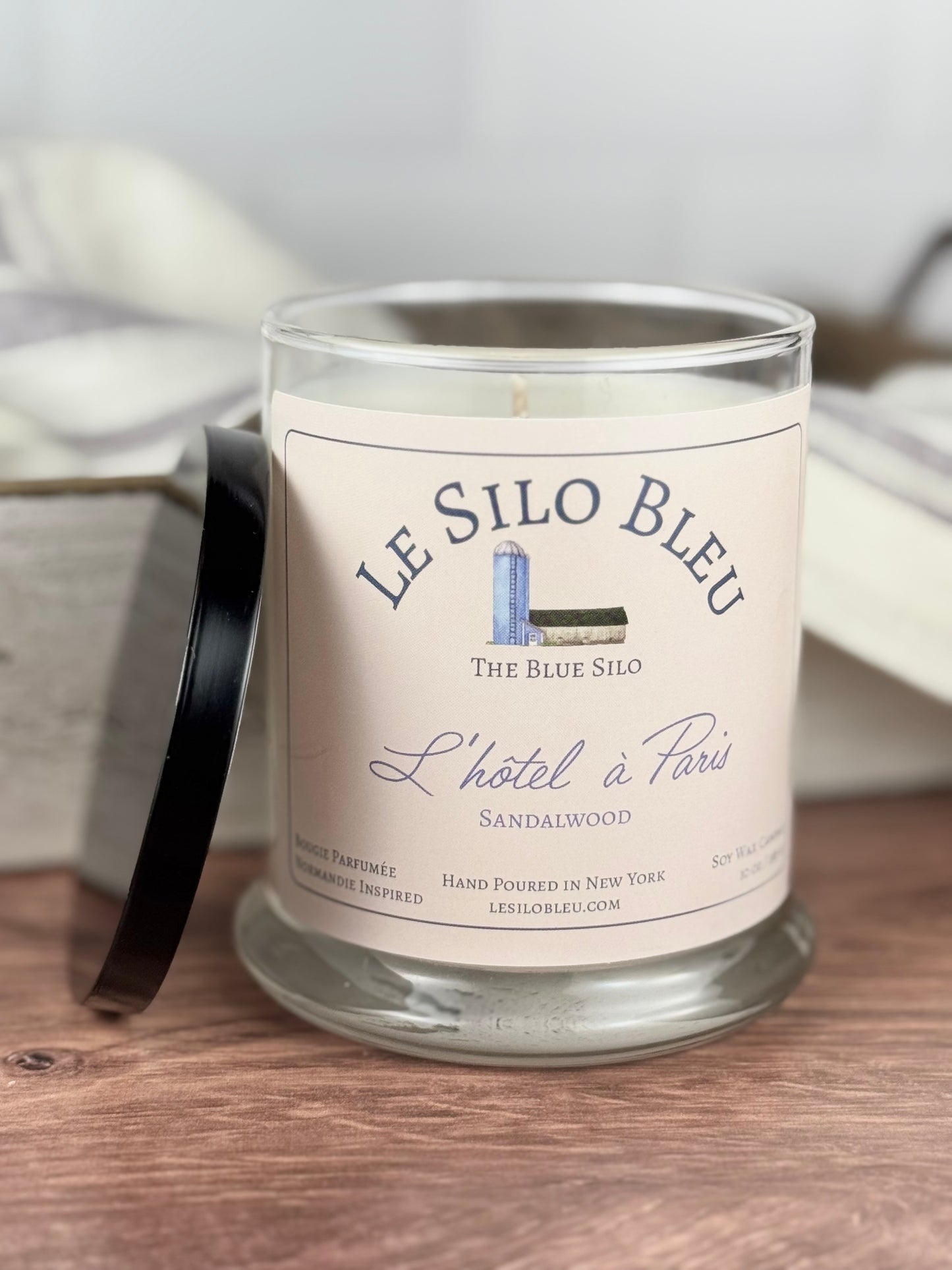 A large handpoured unlit soy wax candle in a glass jar labeled "L'Hotel a Paris- Sandalwood" sitting on a wooden table top with a black lid next to the jar. In the background you can see a white linen towel with lavender stripes in an antique box with a metal handle. 