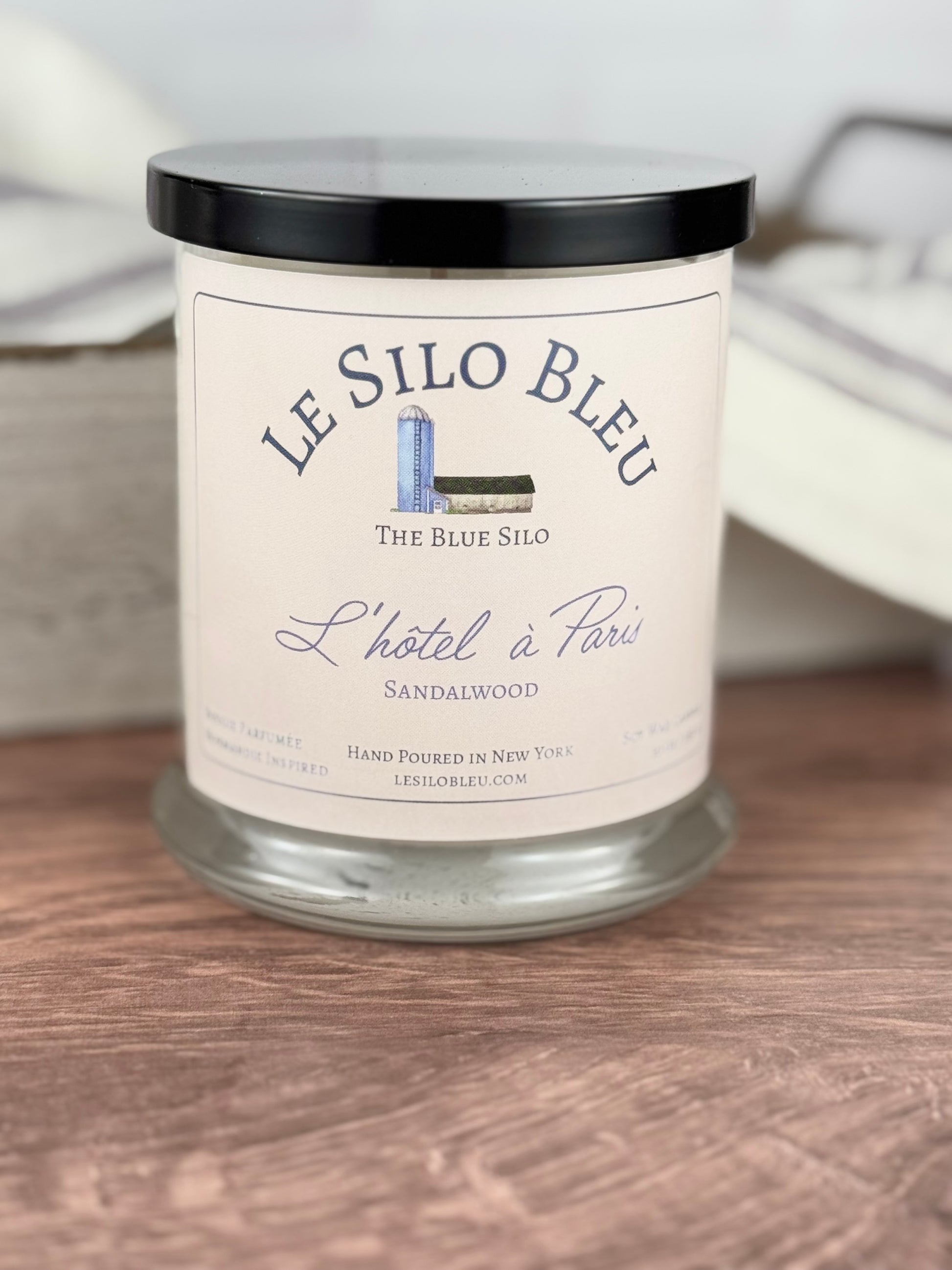 A large handpoured unlit soy wax candle in a glass jar labeled "L'hotel a Paris - Sandalwood" sitting on a wooden table top with a black lid on the jar. In the background you can see a white linen towel with lavender stripes in an antique box with a metal handle. 