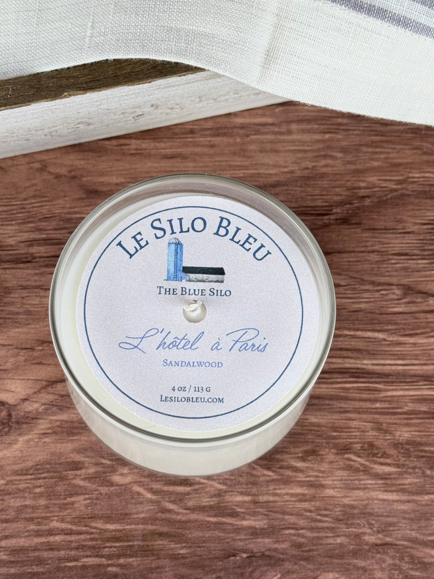 A small handpoured unlit soy wax candle in a glass jar labeled "L'hotel a Paris - Sandalwood" sitting on a wooden table top. In the background you can see a white linen towel with lavender stripes in an antique box.