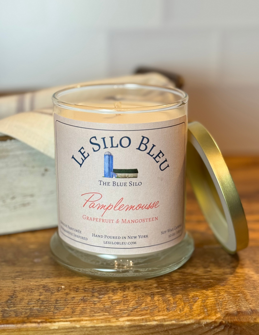 Pamplemousee, a grapefruit and mangosteen scented candle with gold lid to the side. Candle is sitting on top of a wood table with an antique box and linen cloth in the background.