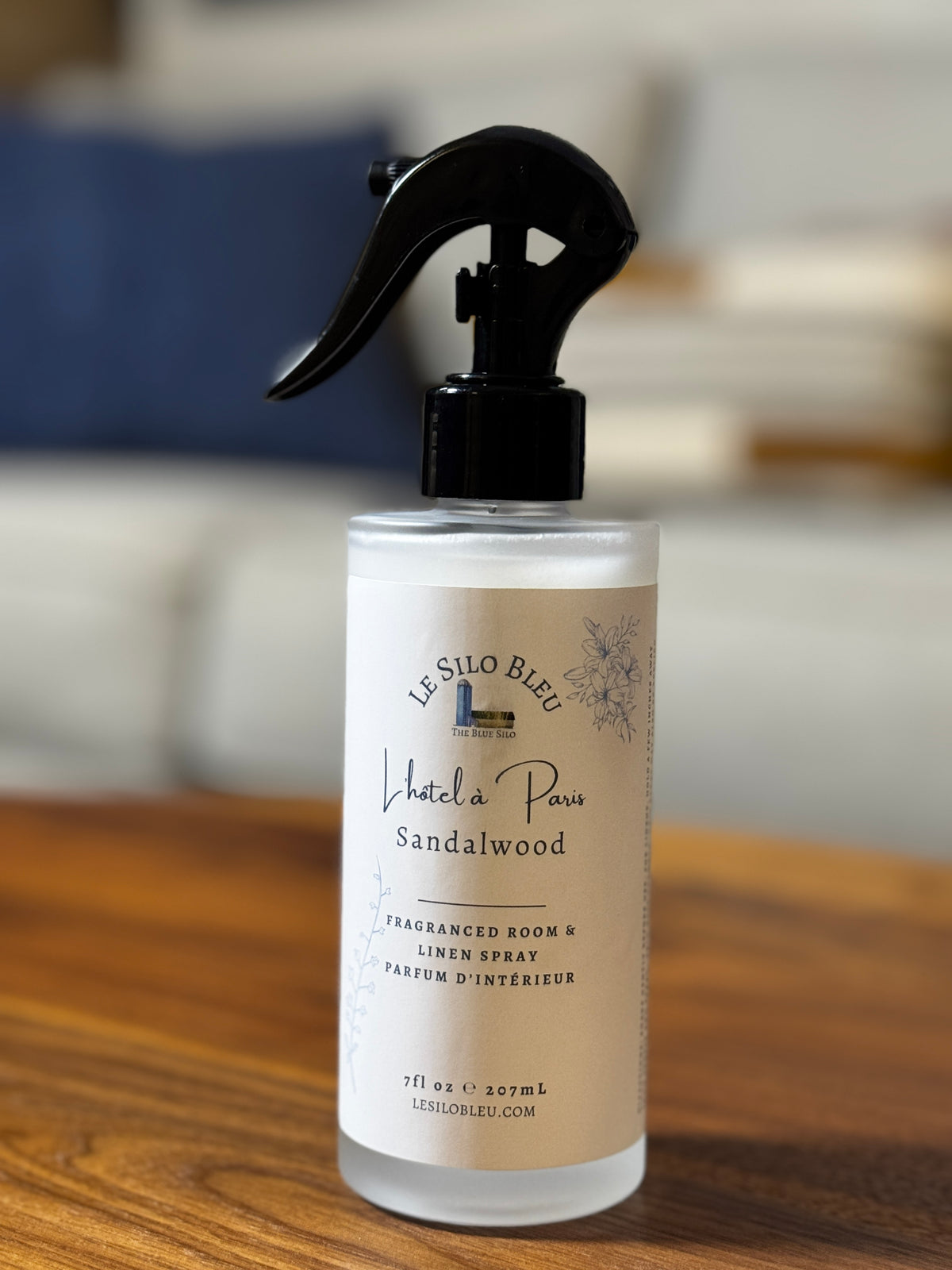 A bottle of Le Silo Bleu 'Hotel in Paris' Sandalwood Fraganced Room & Linen Spray, featuring a black trigger spray top and a cream-colored label, resting on a wooden table with a blurred indoor background.