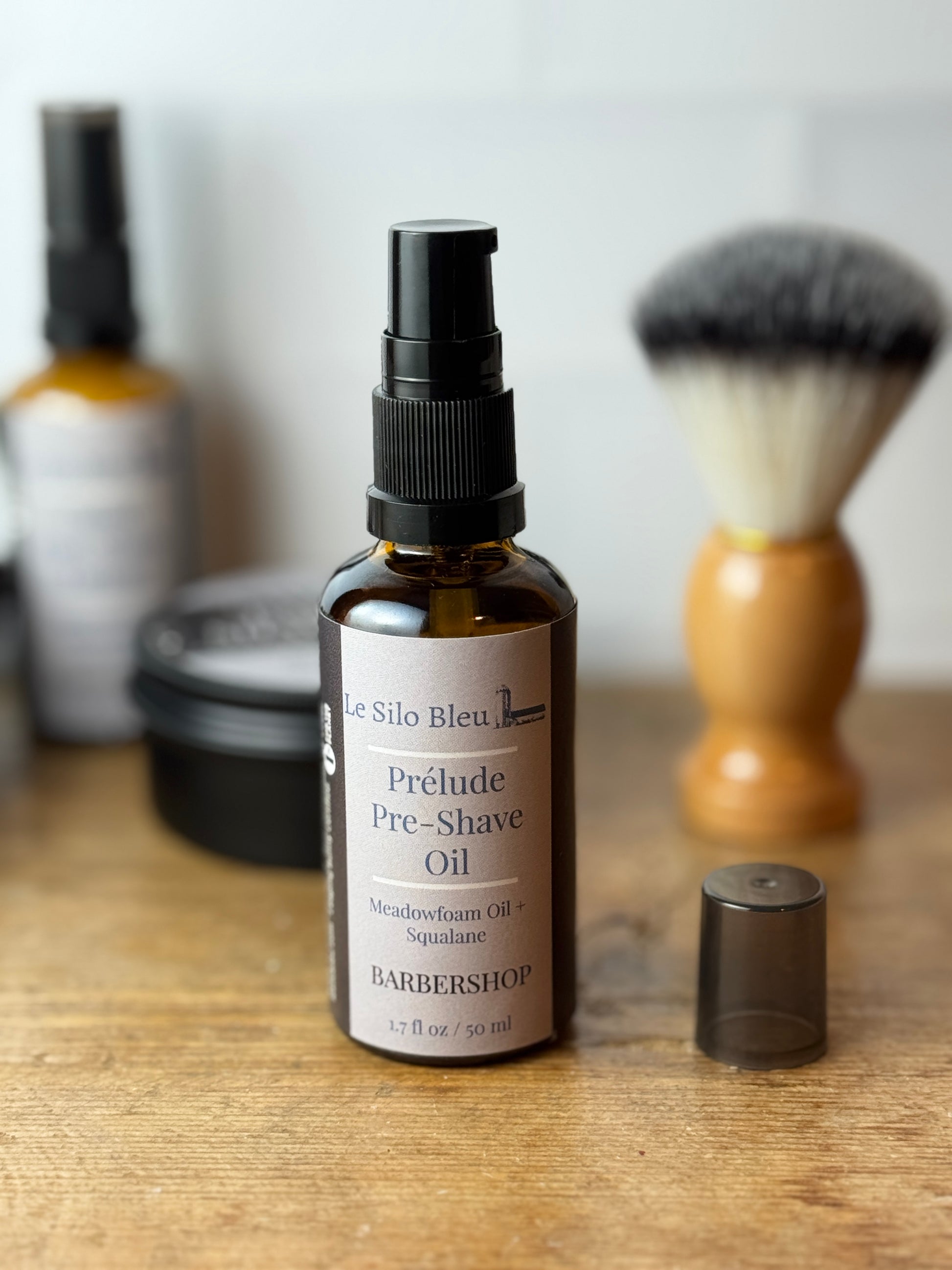 A bottle of Le Silo Bleu Prélude Pre-Shave Oil with its cap removed, placed on a wooden surface with a shaving brush and soap dish blurred in the background.
