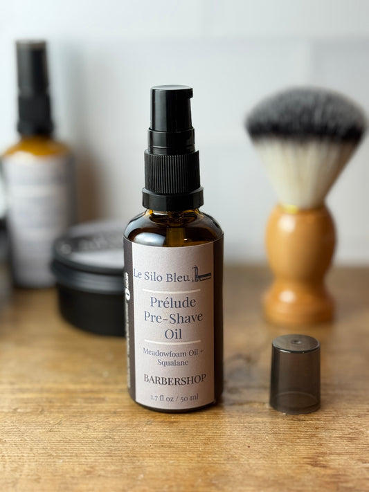 A bottle of Le Silo Bleu Prélude Pre-Shave Oil with its cap removed, placed on a wooden surface with a shaving brush and soap dish blurred in the background.
