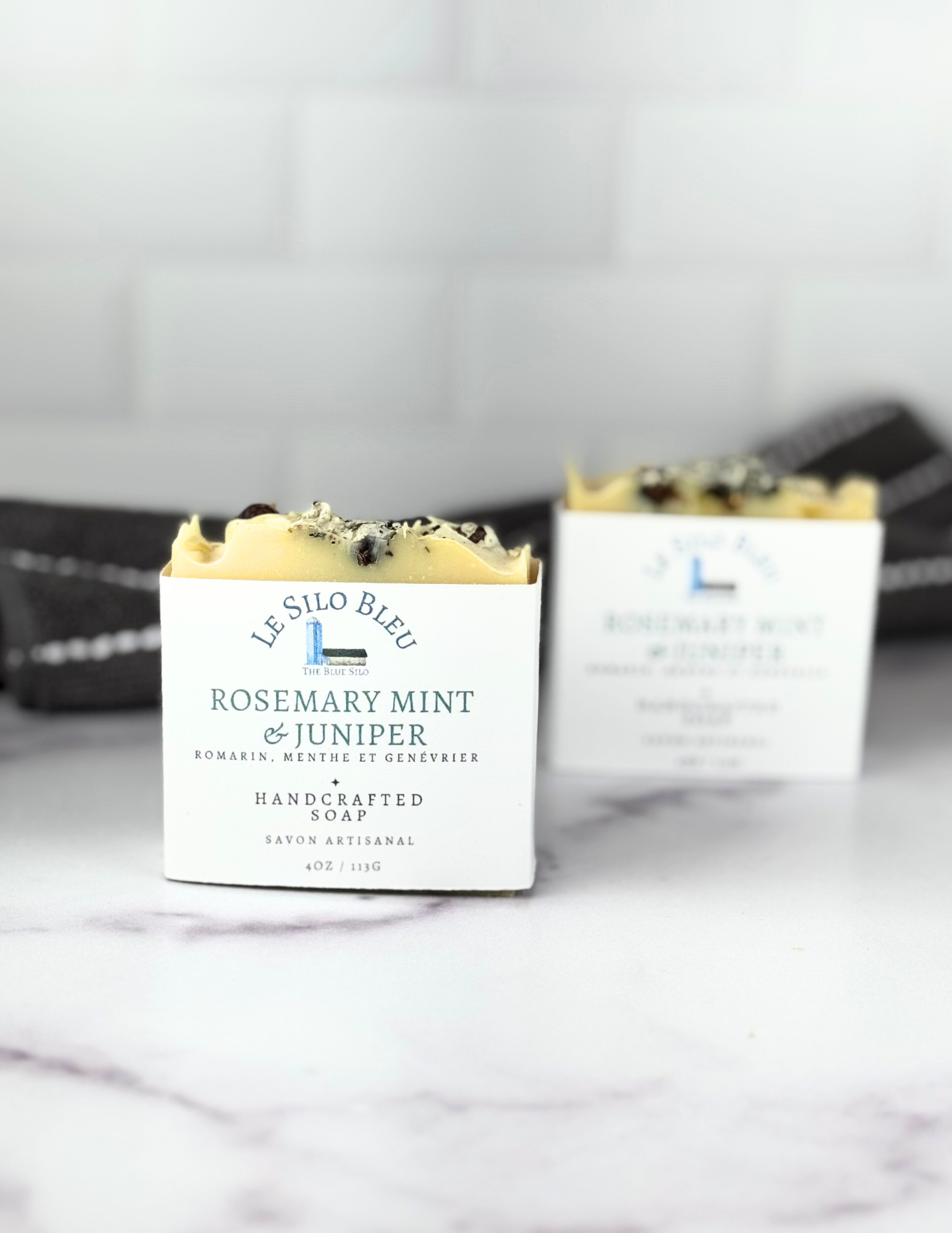 Two bars of labeled soap sit on a white marble tabletop with a grey and white stripped towel in the background. 