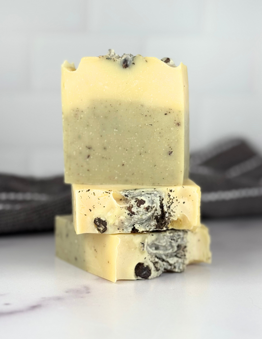 Three Bars of Rosemary Mint and Juniper Berry Soap sitting on a white marble tabletop with a grey and white stripped towel in the background. The soap is green and yellow and speckled with black and brown herbaceous pieces. The top has juniper berries visible. 