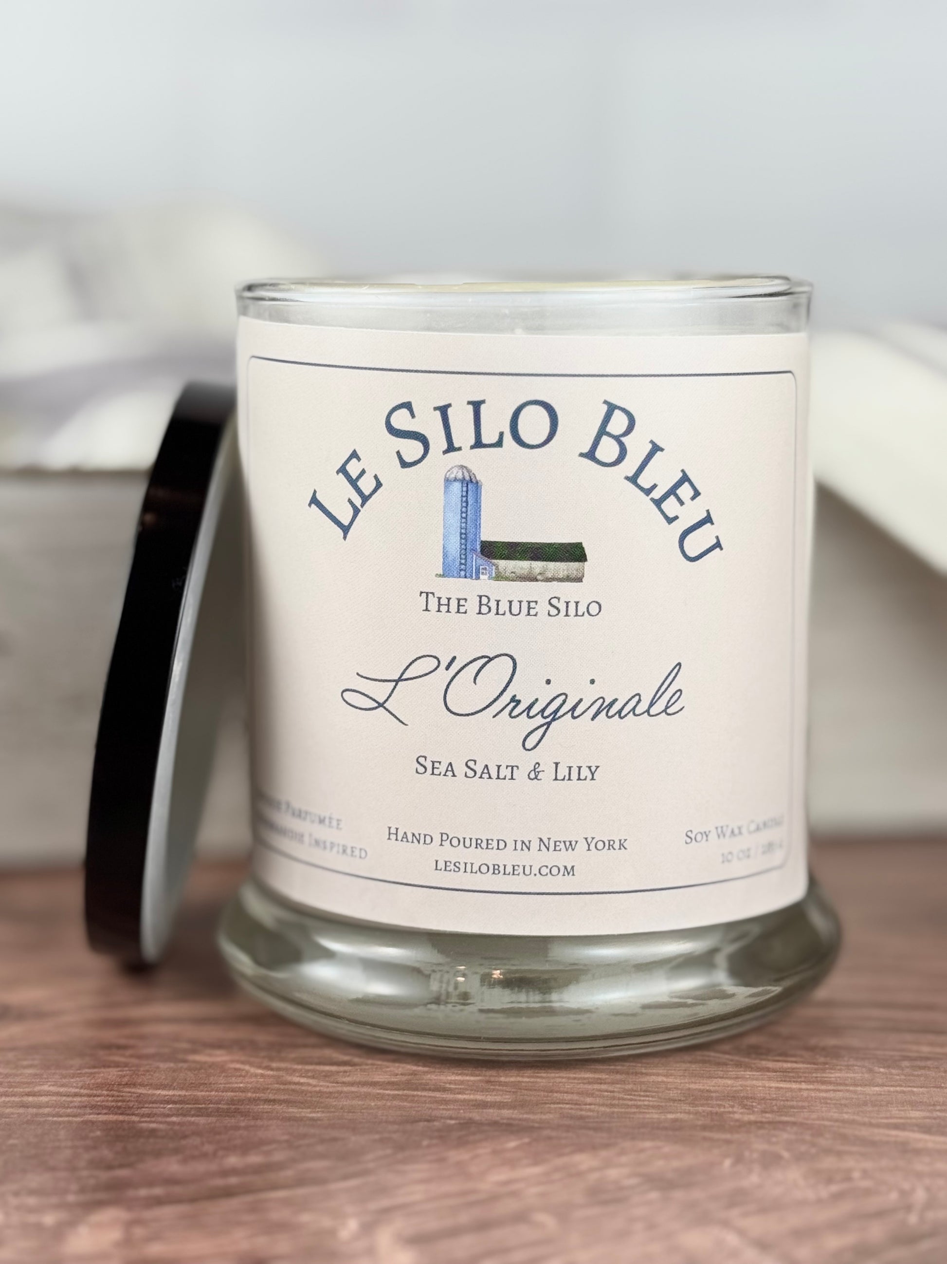 A hand poured unlit soy wax candle in a glass jar labeled " L'Originale-Sea Salt & Lily " sitting on a wooden tabletop with a black lid next to the jar. In the background there is a white linen towel with lavender stripes in an antique box with a metal handle. 