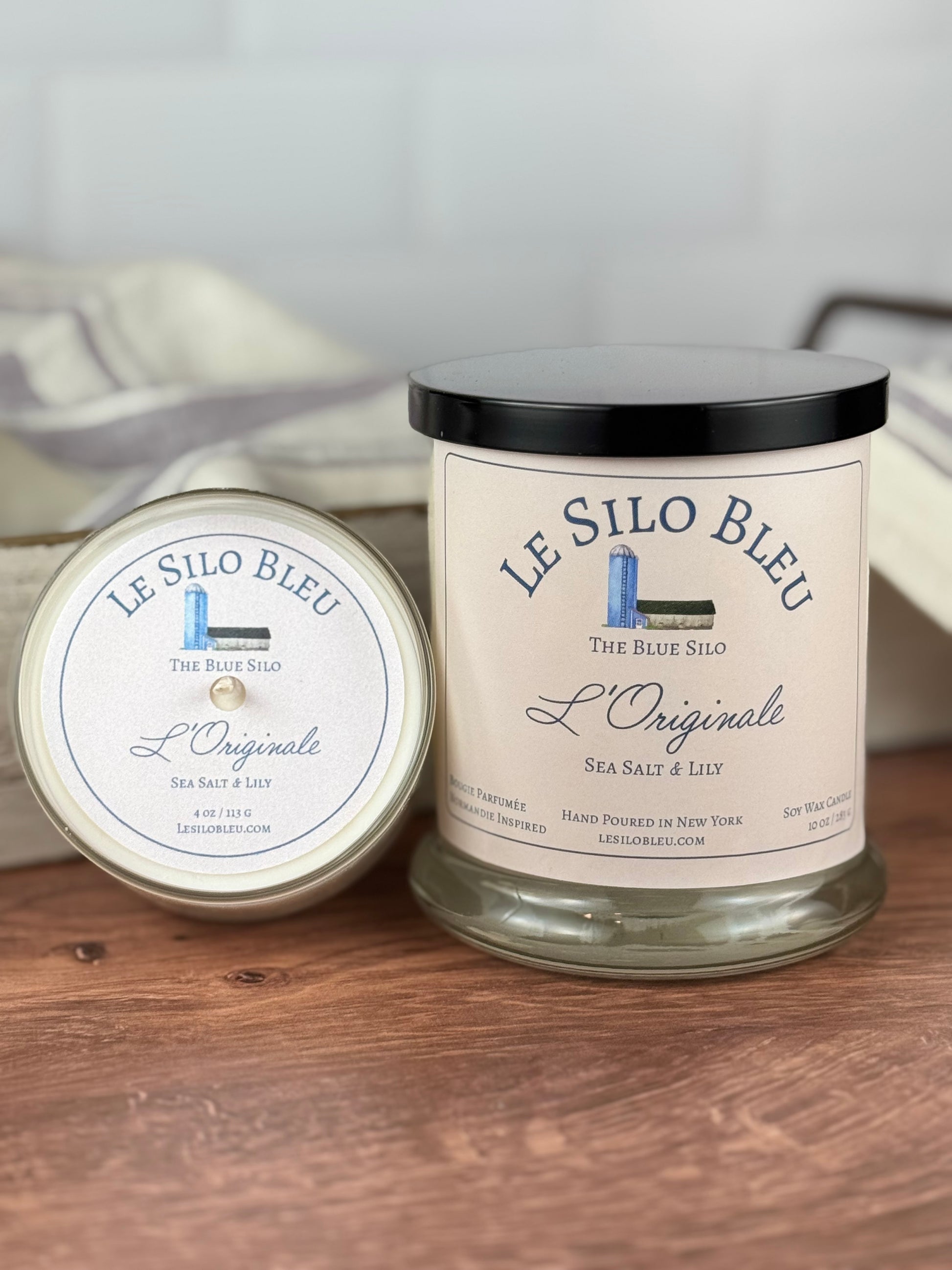 Twp sizes of hand poured unlit soy wax candles in a glass jars labeled " L'Originale-Sea Salt & Lily " sitting on a wooden tabletop with a black lid on the jar. In the background there is a white linen towel with lavender stripes in an antique box with a metal handle. 