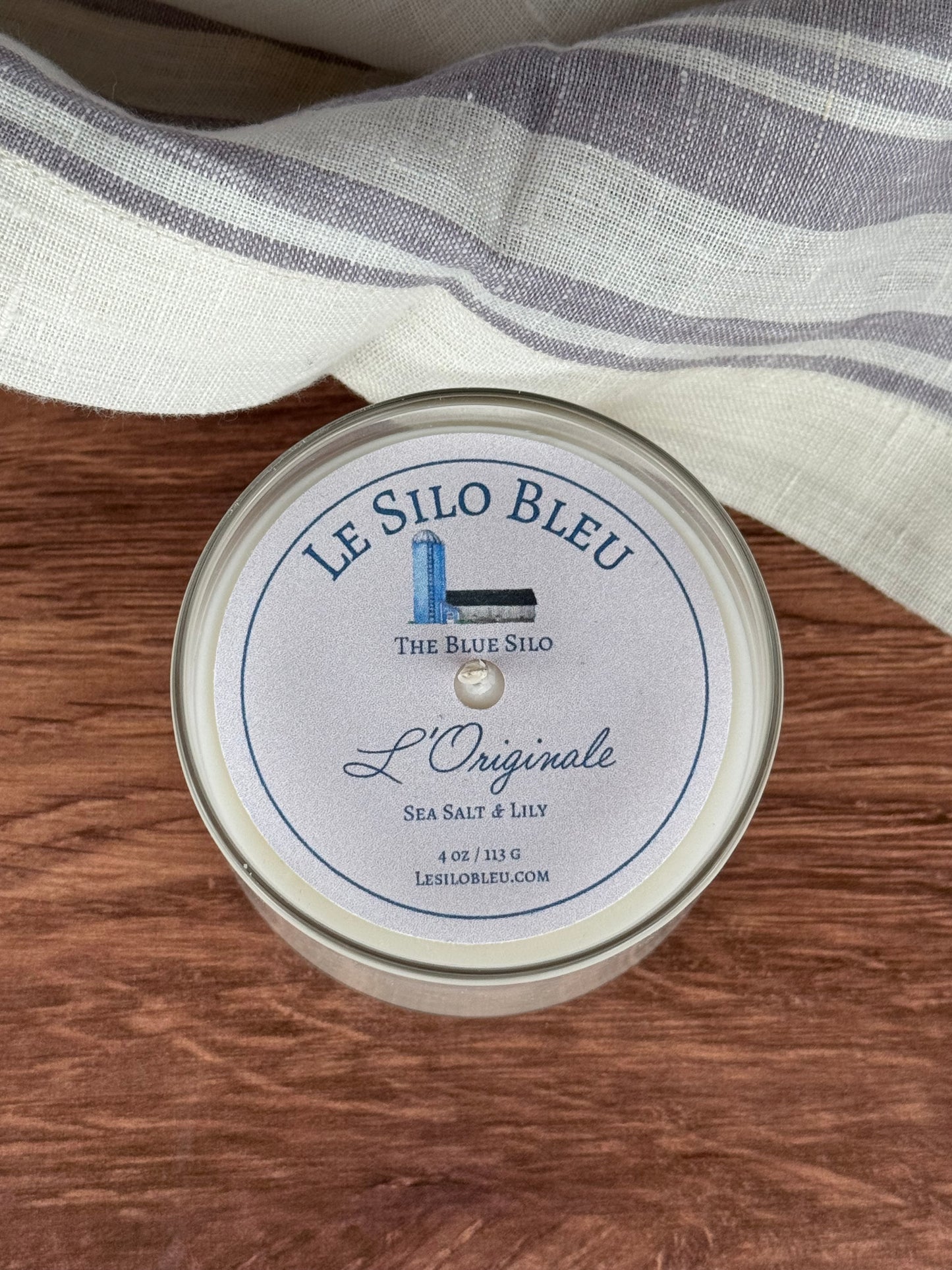 Small sized hand poured unlit soy wax candle in a glass jars labeled " L'Originale-Sea Salt & Lily " sitting on a wooden tabletop. In the background there is a white linen towel with lavender stripes in an antique box.