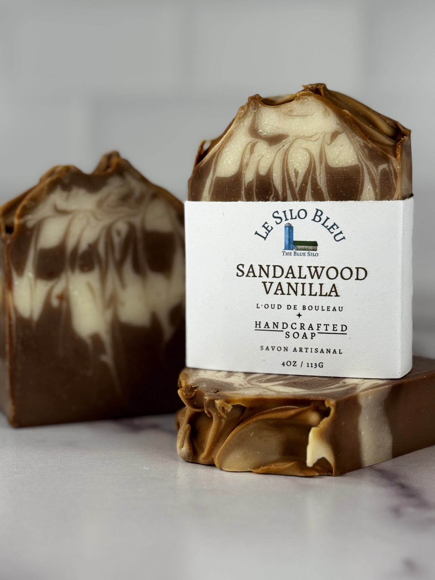 Two handmade artisanal soaps with a marbled brown and cream pattern, featuring a white paper label on the front soap that reads 'Le Silo Bleu Sandalwood Vanilla'.