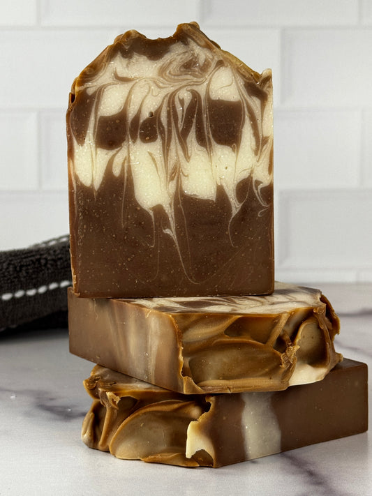 A close-up of a handmade soap bar with a swirled pattern of dark brown and cream, shown with two additional pieces stacked behind it on a light marble-like surface.