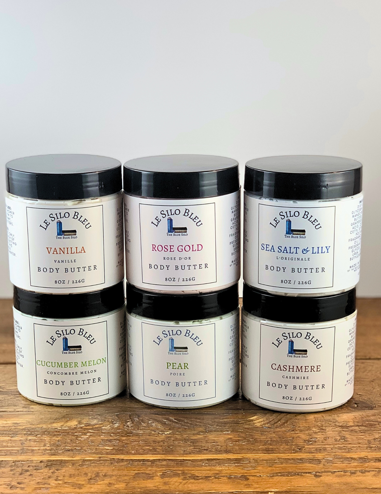 six jars of body butter stacked in two rows on a wooden table with a white background.