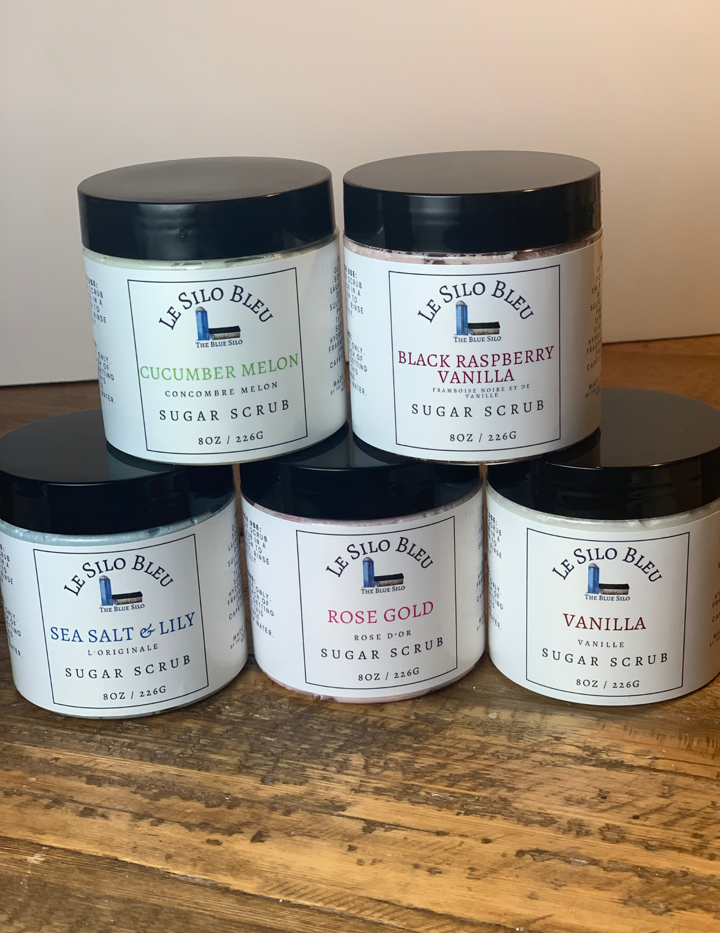 Stacks of sugar scrubs with new labels on a wooden tabletop with a white background.The jars have black lids.