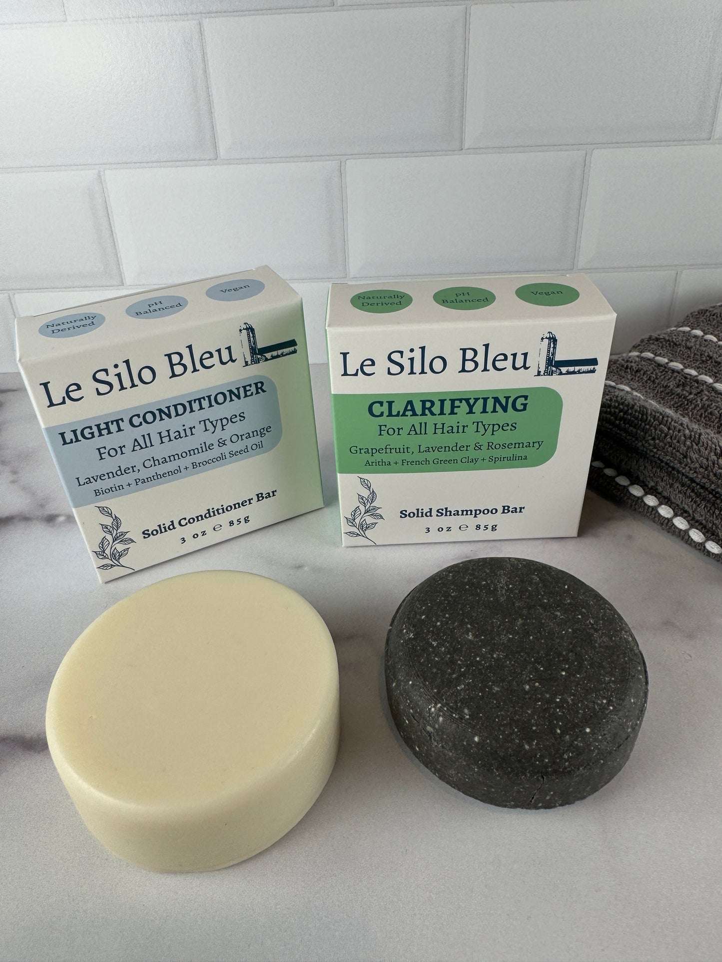 Clarifying shampoo and light conditioner solid bars with their box and a grey towel on a marble surface with a white tiled wall in the background