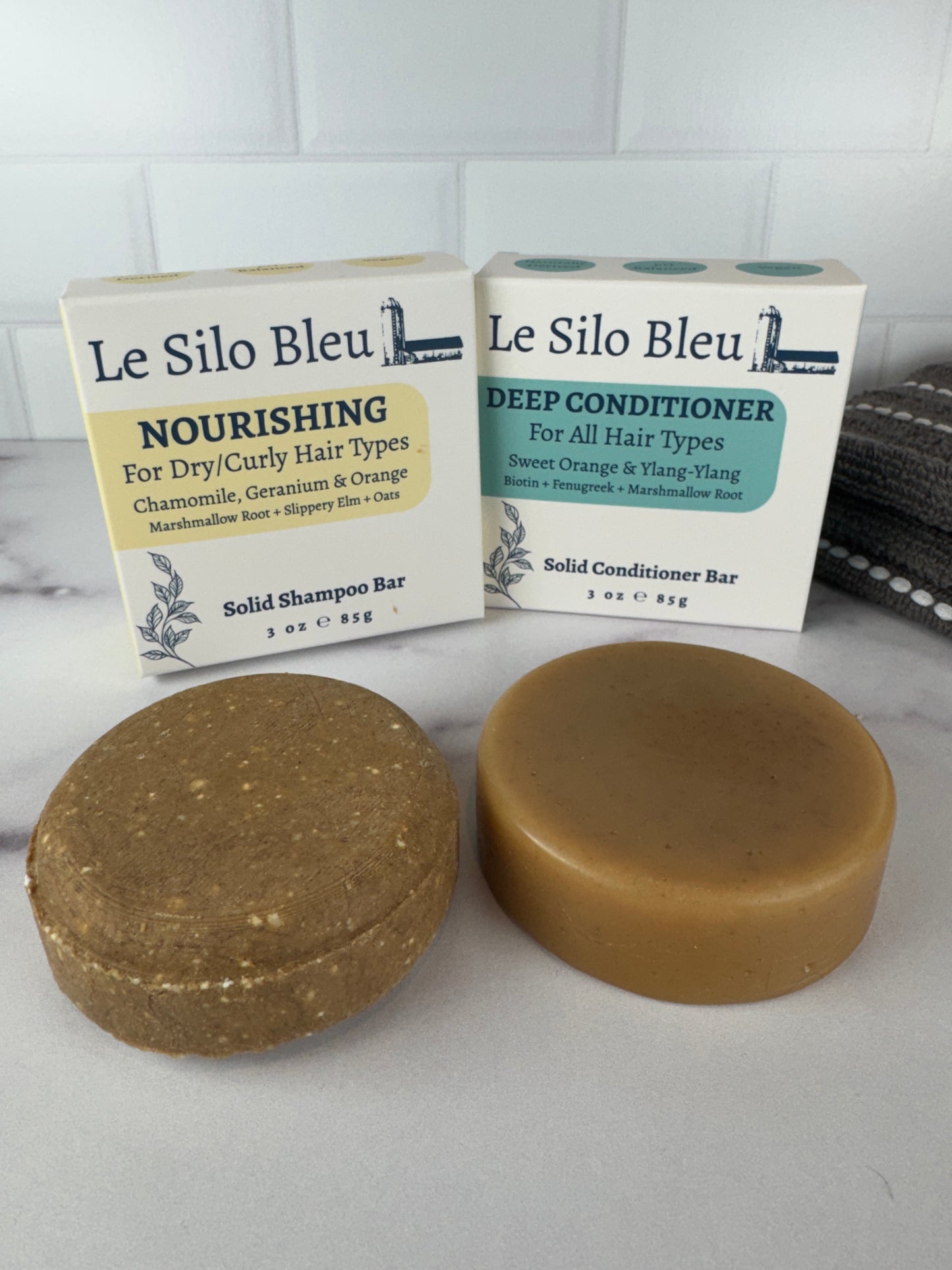 Two solid shampoo and conditioner soap bars and their packaging on a white surface with a grey towel in the background next to white tiled wall.