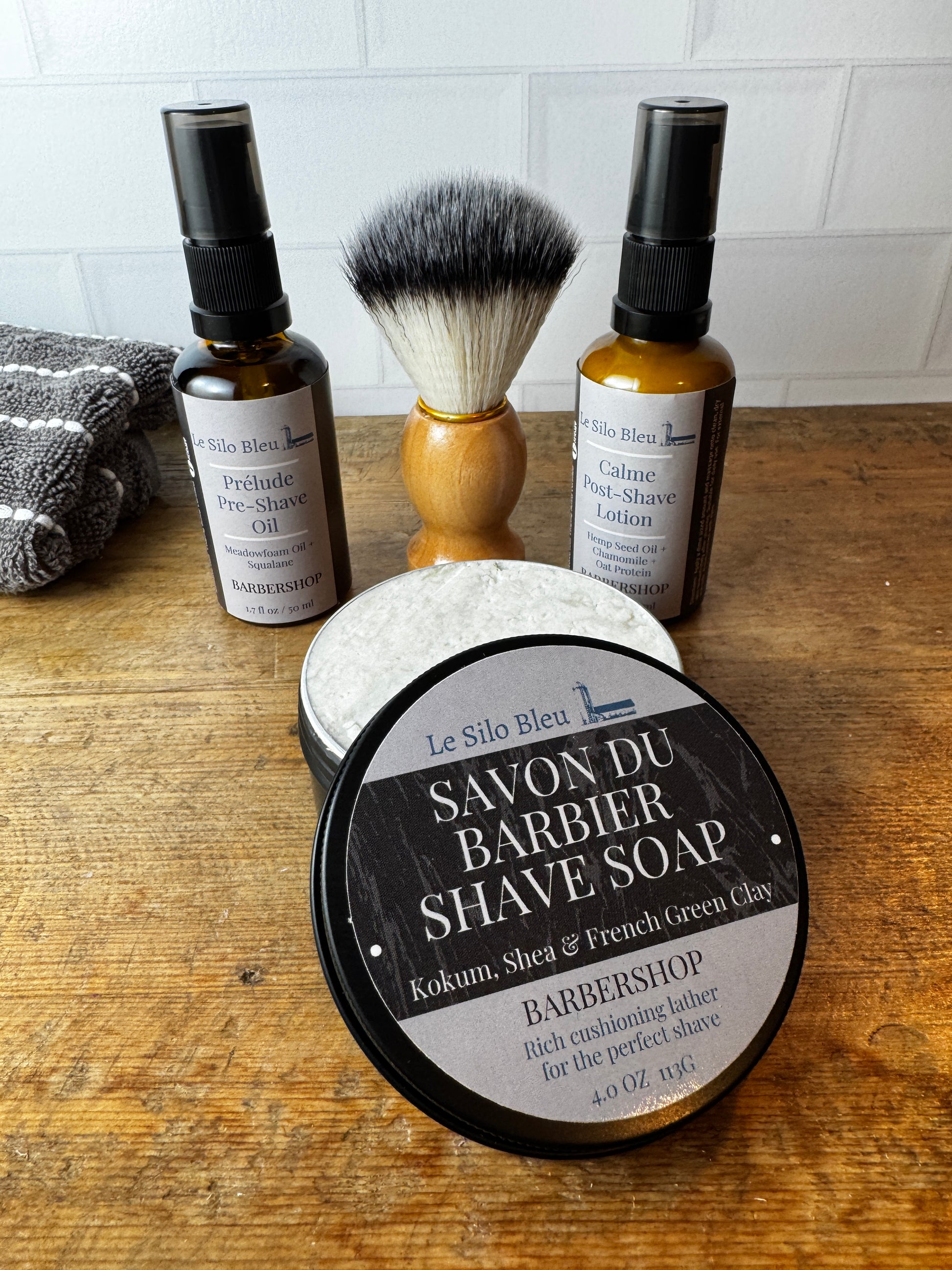 A barbershop grooming set on a wooden table, featuring a round tin of Le Silo Bleu Savon du Barbier Shave Soap in the Kokum, Shea & French Green Clay scent, alongside a shaving brush, pre-shave oil, and calamine lotion.