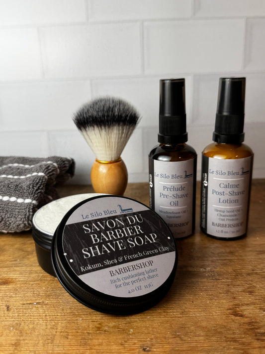 A grooming set arranged on a wooden surface against a white subway tile wall, including two amber bottles with black caps, a shaving brush with black and white bristles and a light wood handle, a black tin of Ultra Shave Soap, and a gray towel.