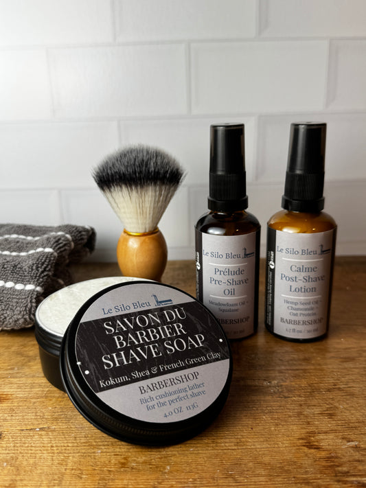 Le Silo Bleu Barbier Shop shaving set on a wooden table, featuring a shaving soap tin, two amber spray bottles, and a shaving brush against a white subway tile background.