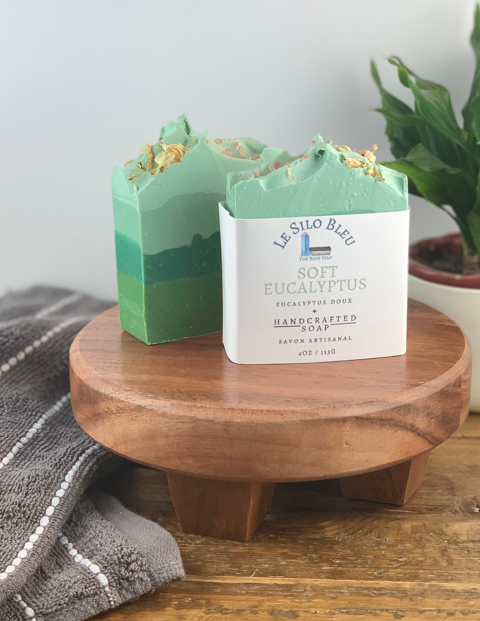 Two bars of Soft Eucalyptus Soap sitting on a wooden platform with one soap wrapped in white labeling and one without. There is a green plant in a white pot in the background and a grey and white striped towel in the foreground. There is a wooden table and white background. 