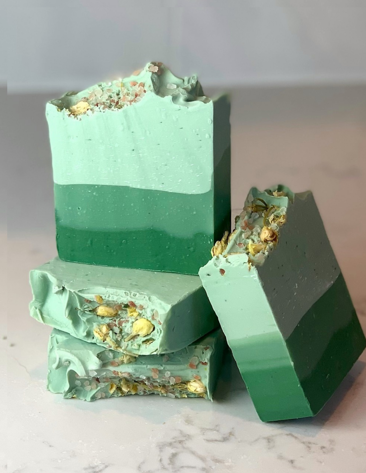 A stack of soft eucalyptus and cotton soap in shades of ombre green colors with jasmine flowers on top all sitting on a a white marble table. 