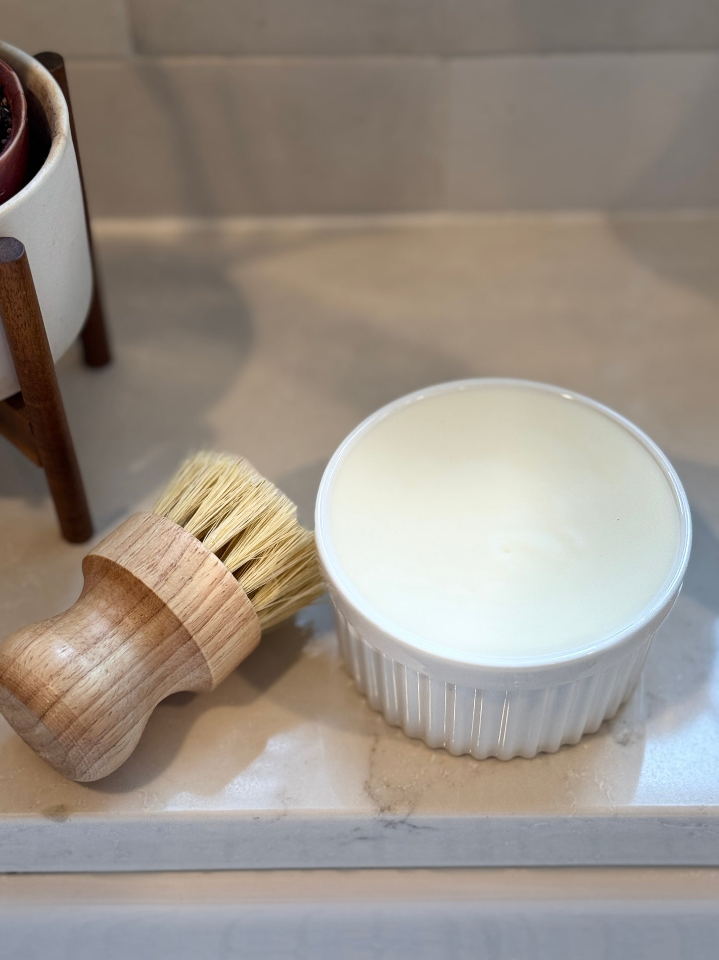 Wooden brush and white ceramic ramekin filled with solid soap on a marble sink top with a plant in the background.
