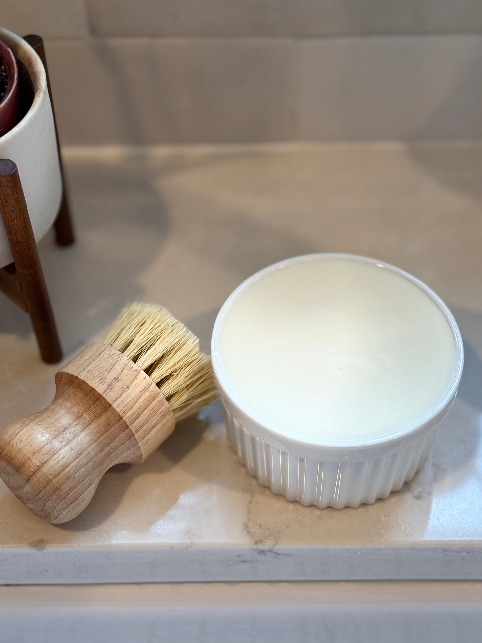 Wooden brush and white ceramic ramekin filled with solid soap on a marble sink top with a plant in the background.