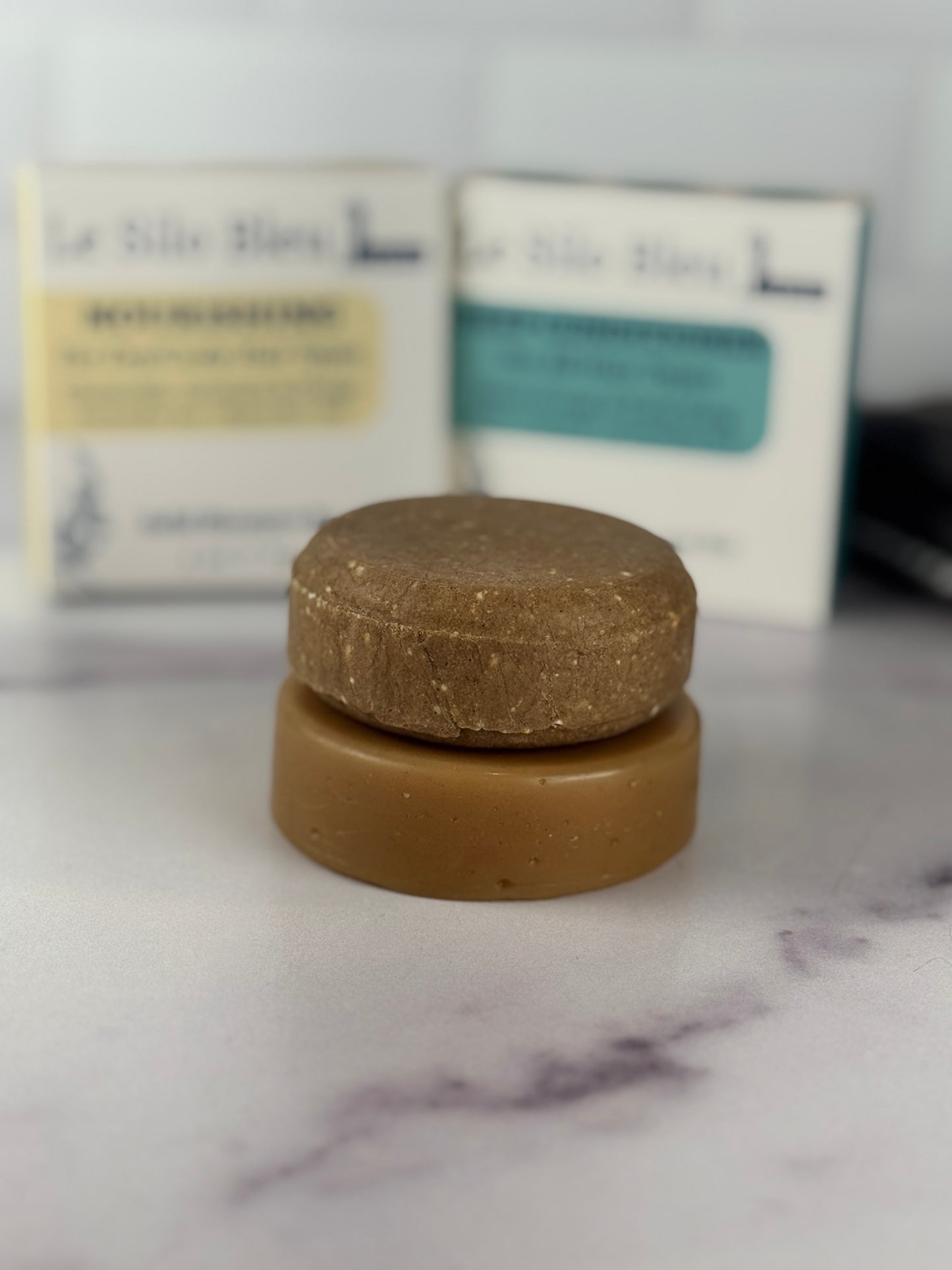 Two solid shampoo and conditioner bars on a marble surface with blurred packaging in the background.