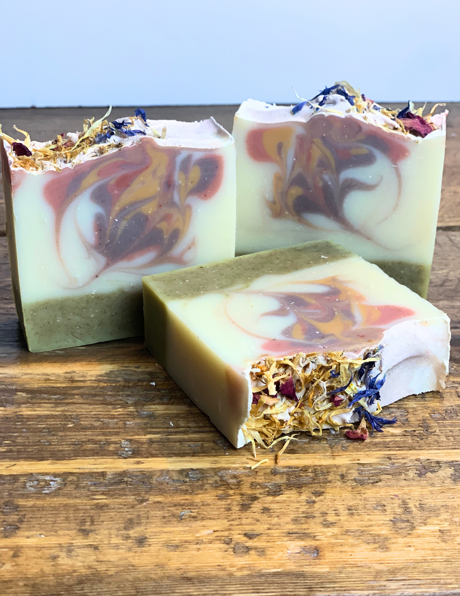 Three Spring Meadow soaps sitting on a wood tabletop with floral botanical petals on top. 