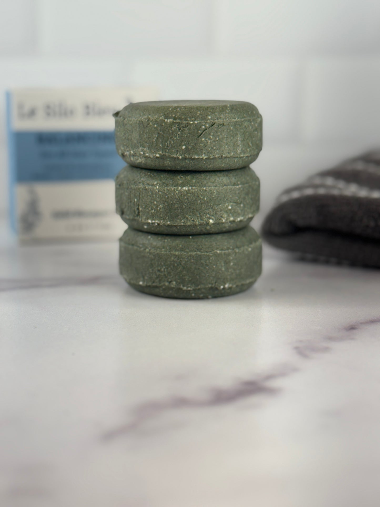 Stack of three dark green Balancing Solid Shampoo bars on a marble surface with a blurred background