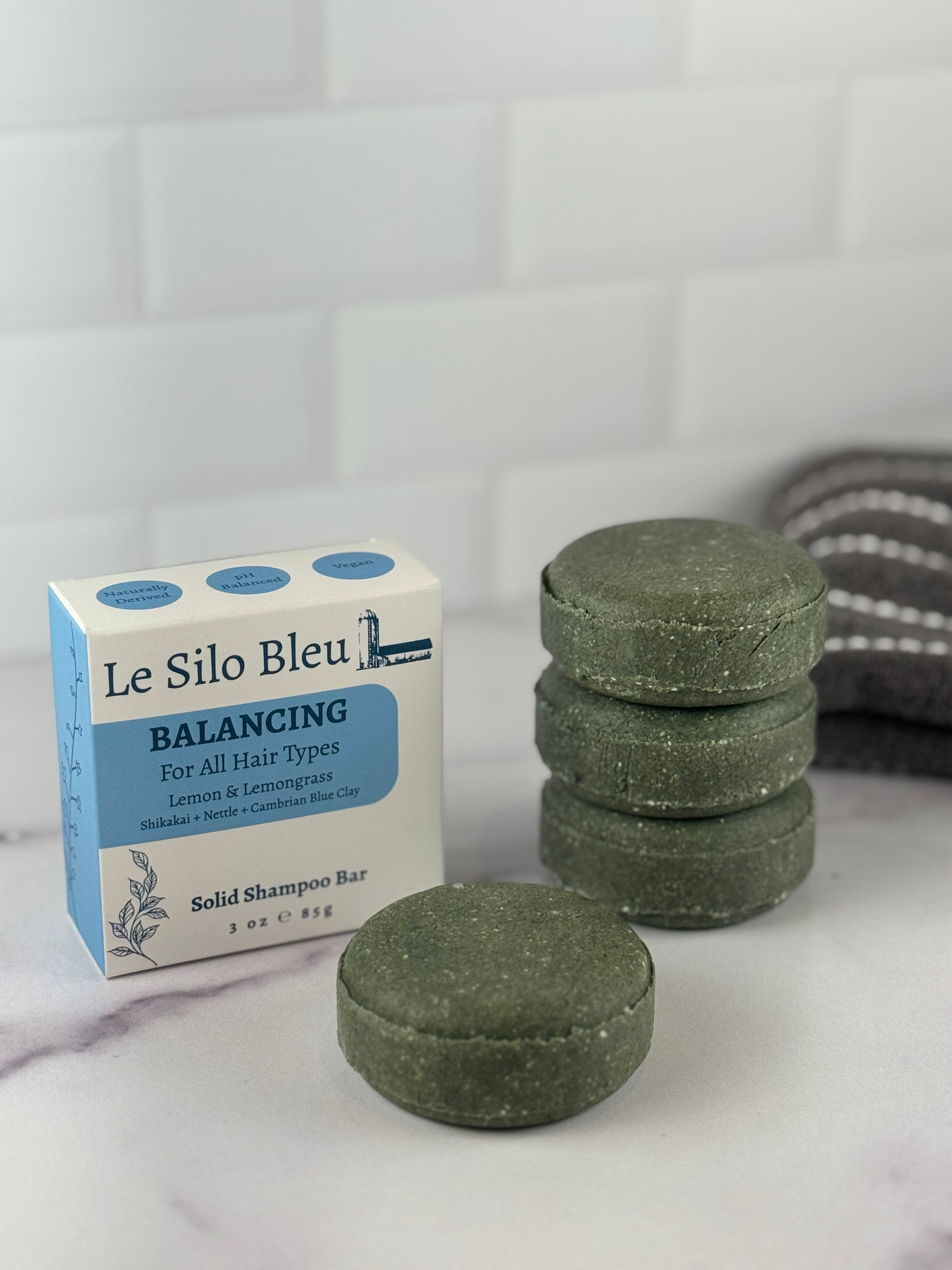 A single and a stack of three dark green Balancing Solid Shampoo Bars and packaging on a marble surface with a white tiled wall background