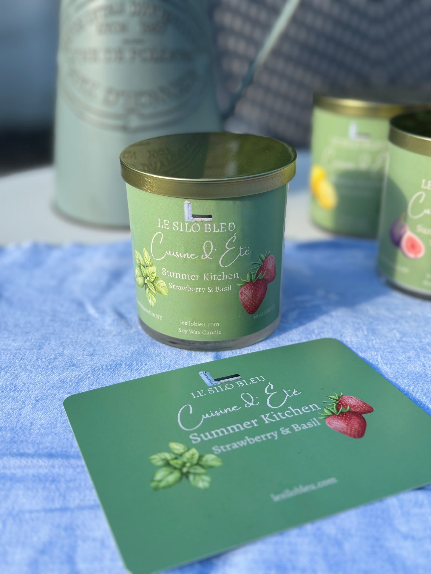 Summer Kitchen Strawberry & Basil Candle with a green label on a blue surface, surrounded by similar candles and a pitcher of flowers in the background. 