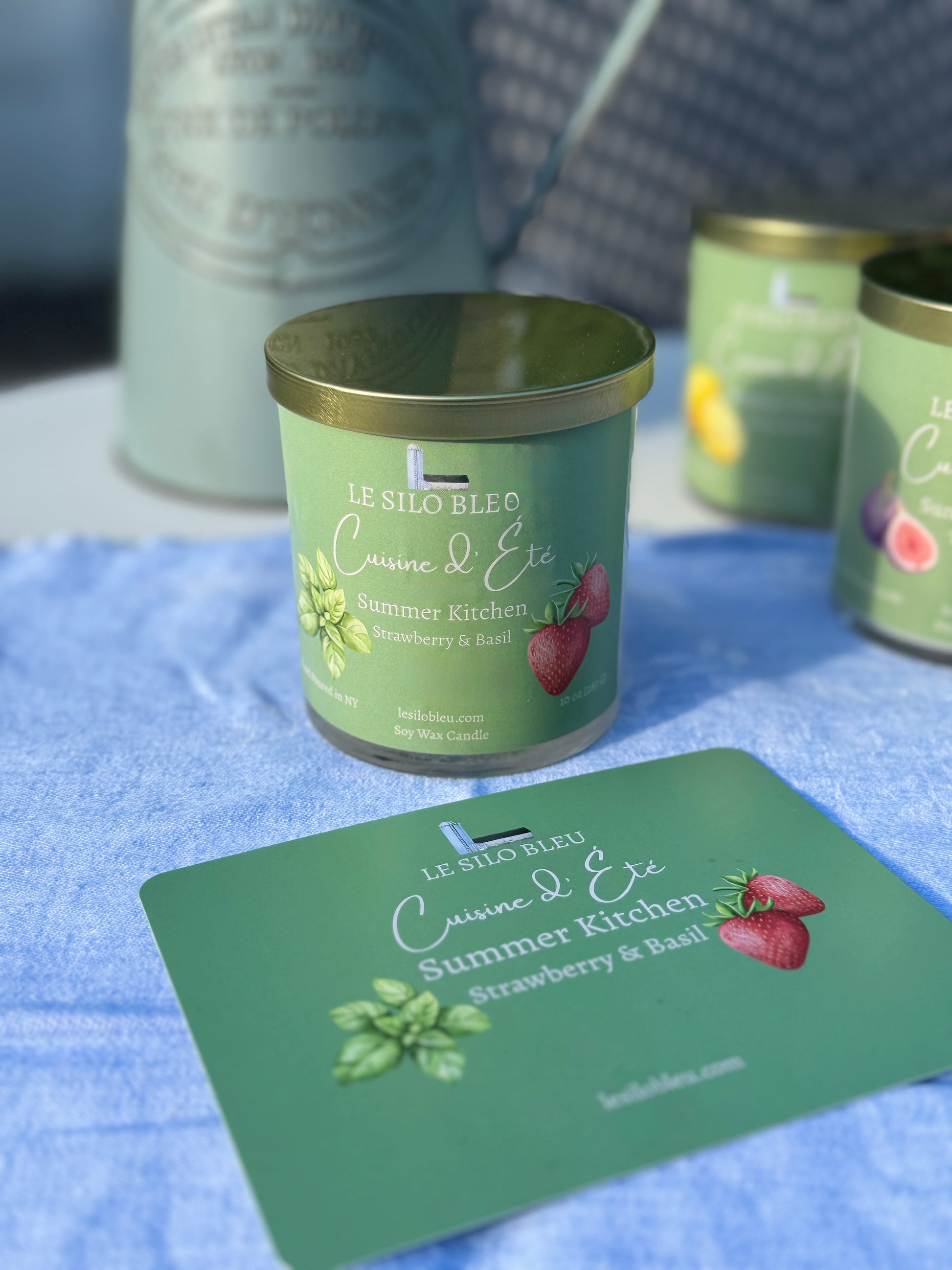 Summer Kitchen Strawberry & Basil Candle with a green label on a blue surface, surrounded by similar candles and a pitcher of flowers in the background. 