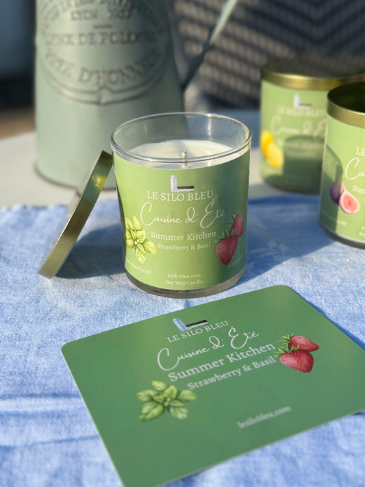 Summer Kitchen Strawberry & Basil Candle with a green label on a blue surface, surrounded by similar candles and a pitcher of flowers in the background. 