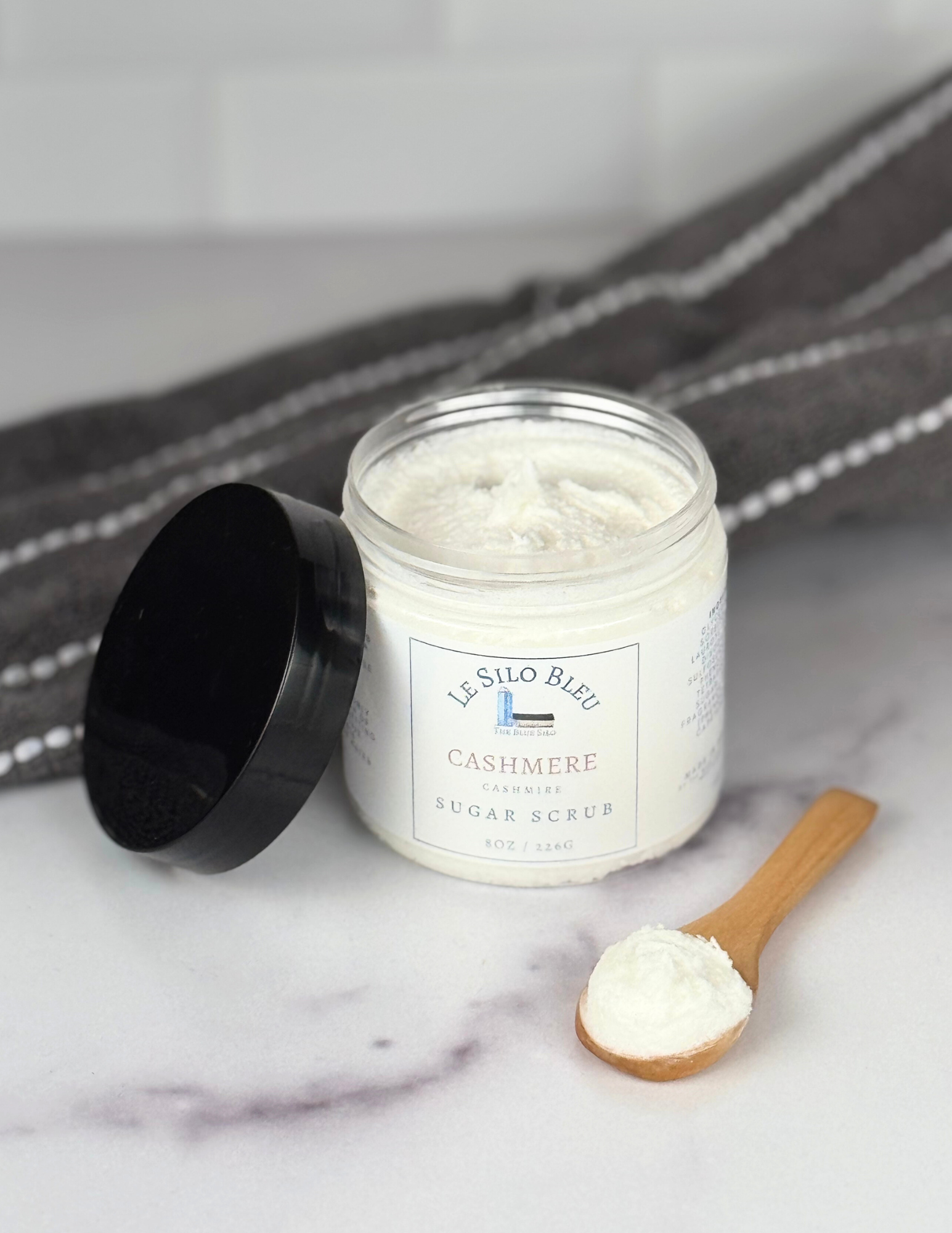 Jar of sugar scrub jar with black lid leaning against the jar. Both are sitting on a white marble table with a grey and white stripped towel in the background. The white labels for these show the logos and names of the scent- Cashmere.  There is also a small wooden spoon with a scoop of sugar scrub on it in the foreground.