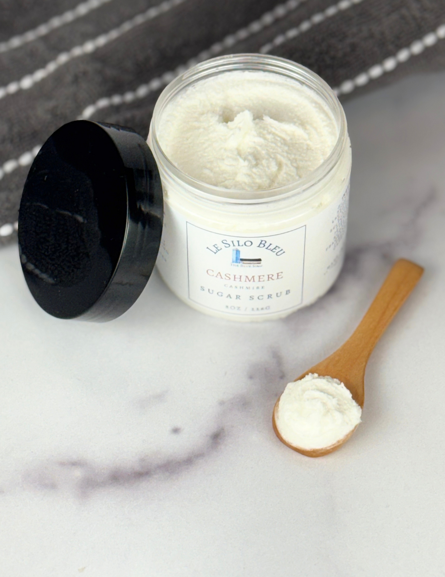 An open jar of Cashmere scented Sugar Scrub with a black lid leaning against the jar sitting on top of a white marble table with a grey and white stripped towel in the background. The label shows the logo and the name of the scent. There is a small wooden scoop spoon with sugar scrub in it on the table as well. 