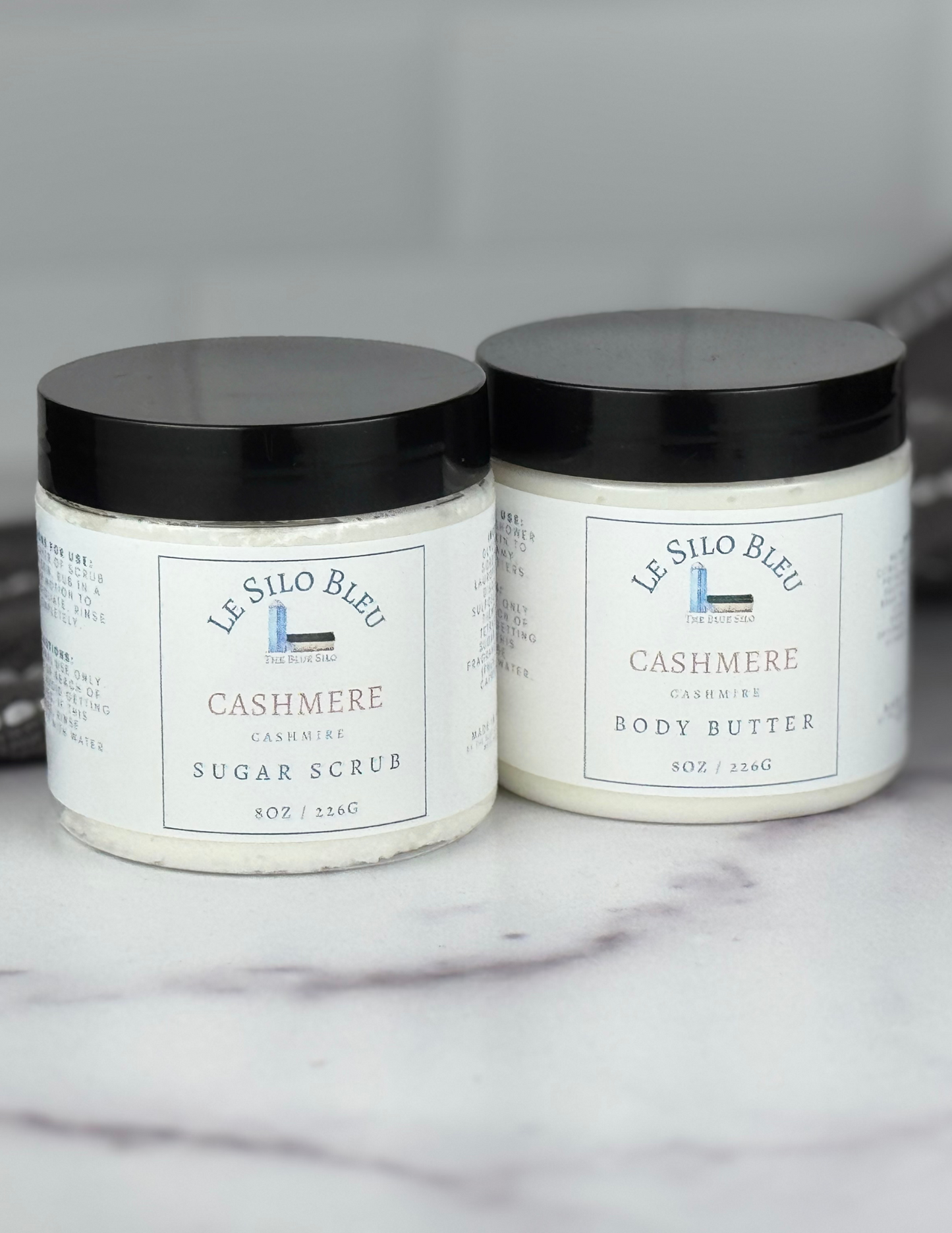 Set of body butter and sugar scrub jars with black lids sitting on a white marble table with a grey and white stripped towel in the background. The white labels for these show the logos and names of the scent- Cashmere. 